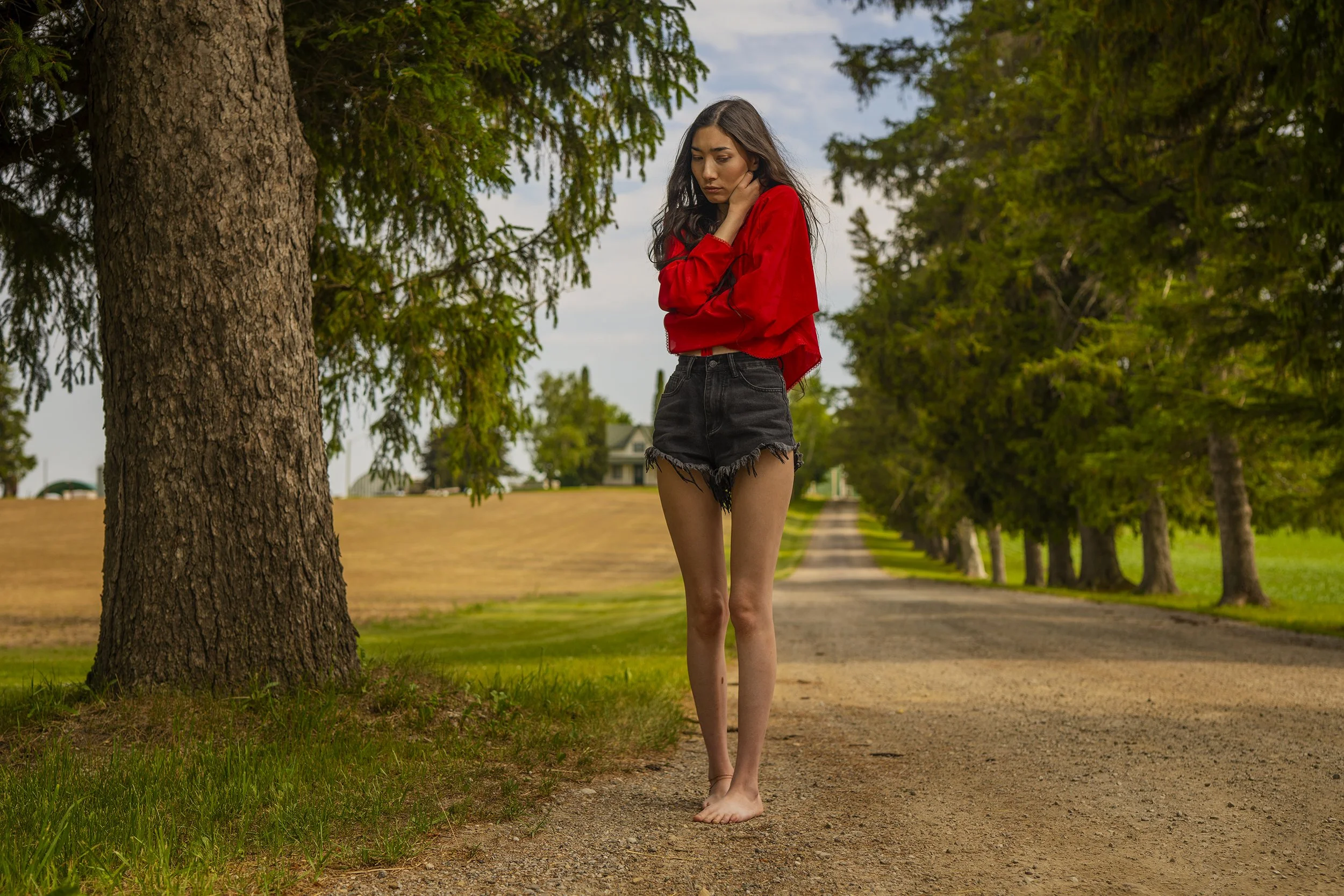 A young woman with long dark hair, wearing a red jacket and black denim shorts, standing barefoot on a dirt pathway in a park lined with tall green trees.