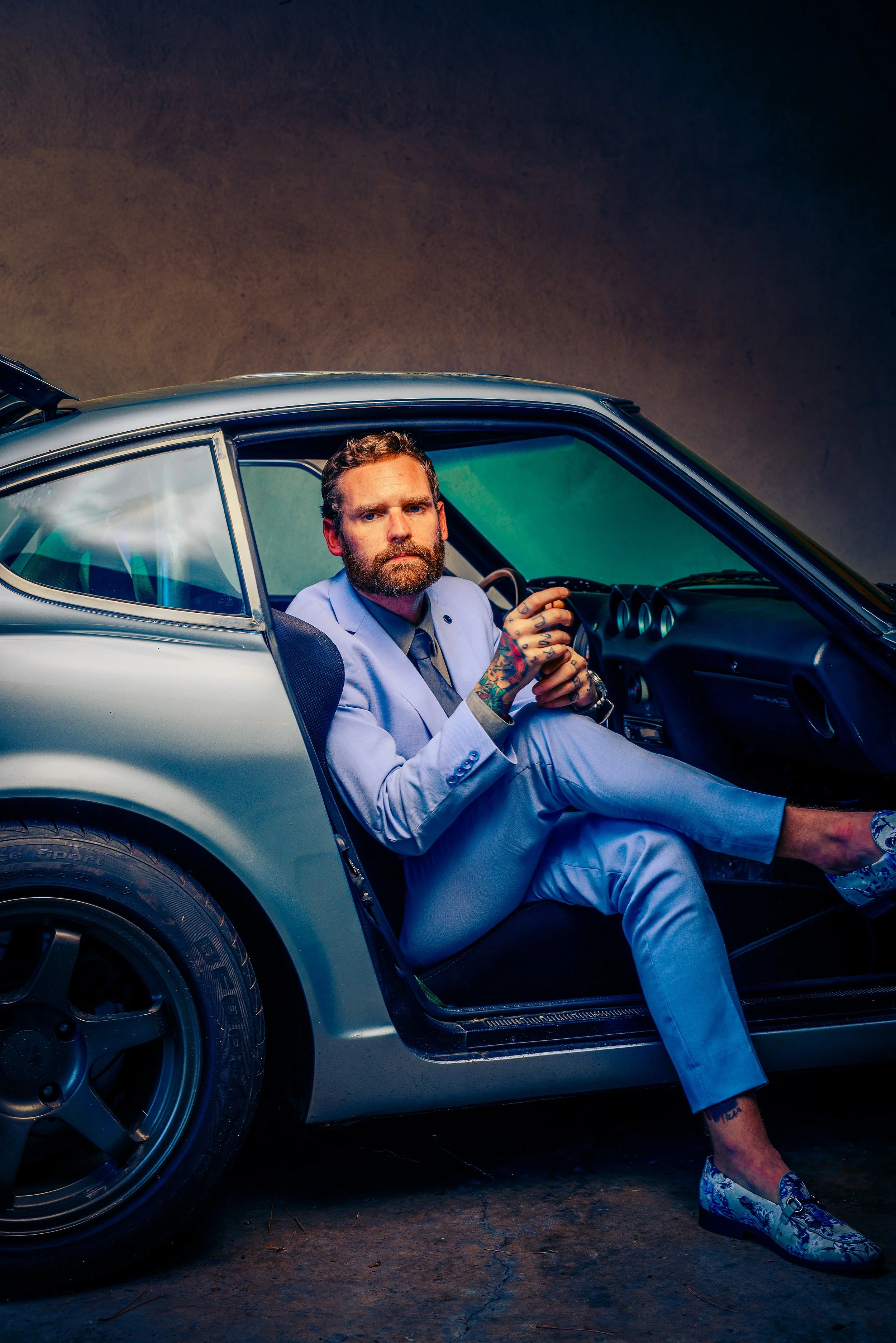 Man with tattoos and beard sitting in a silver sports car, dressed in a light blue suit, with a dark background.