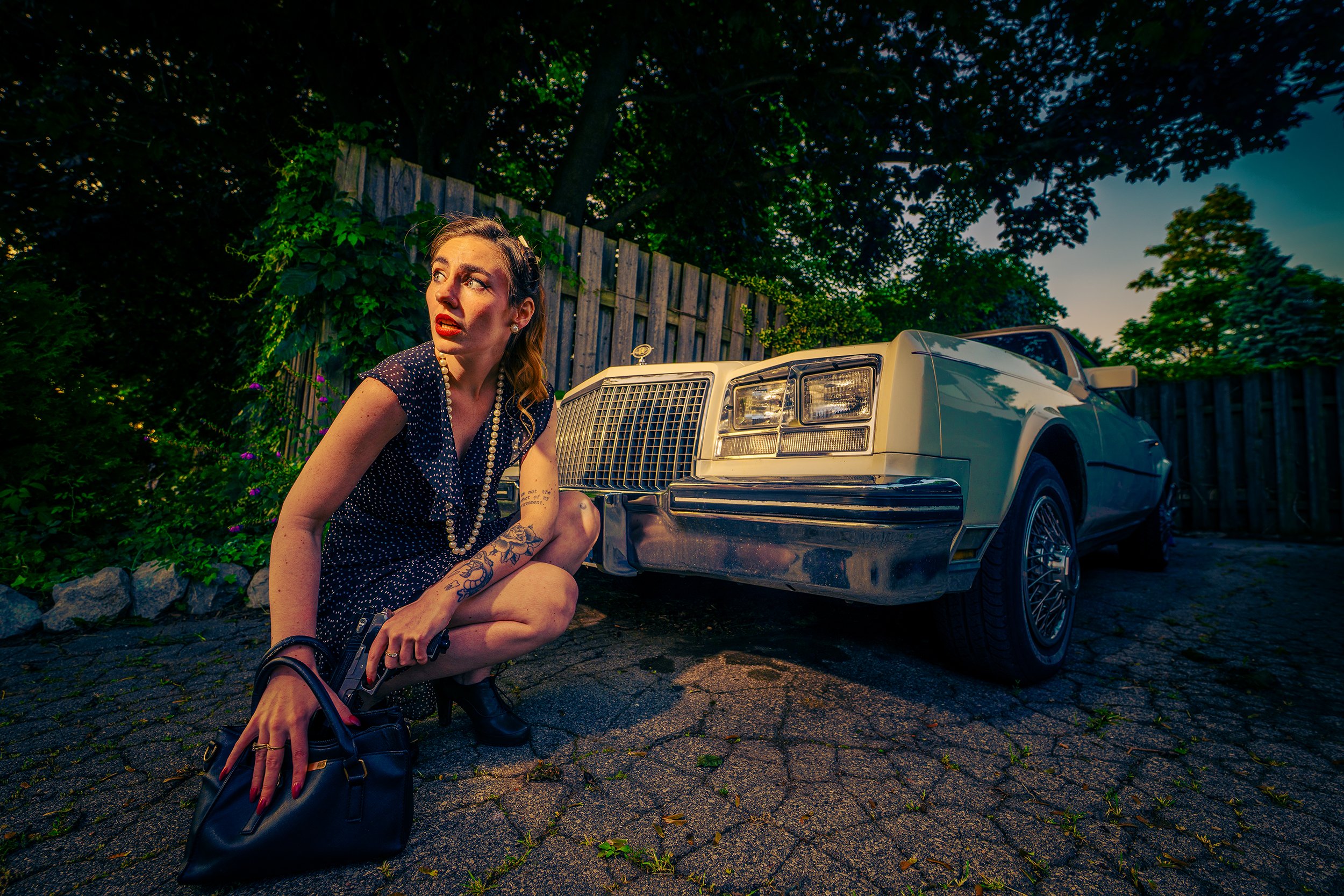 A woman with tattoos crouching on pavement next to a vintage white car, looking to her left with a concerned expression, in a backyard with wooden fence and green trees at dusk.
