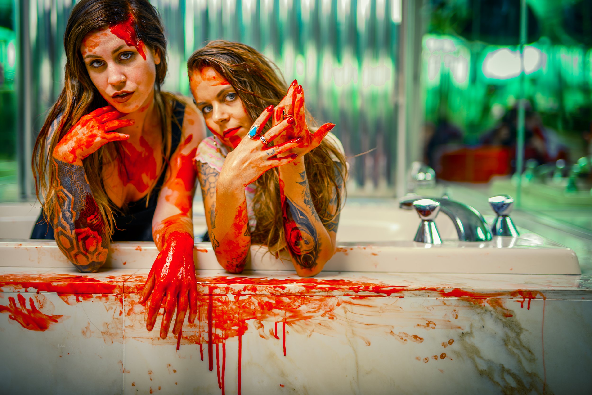Two women covered in fake blood with plastic gloves, posing behind a blood-splattered sink in a room with green and clear plastic walls, created for a horror-themed or theatrical event.