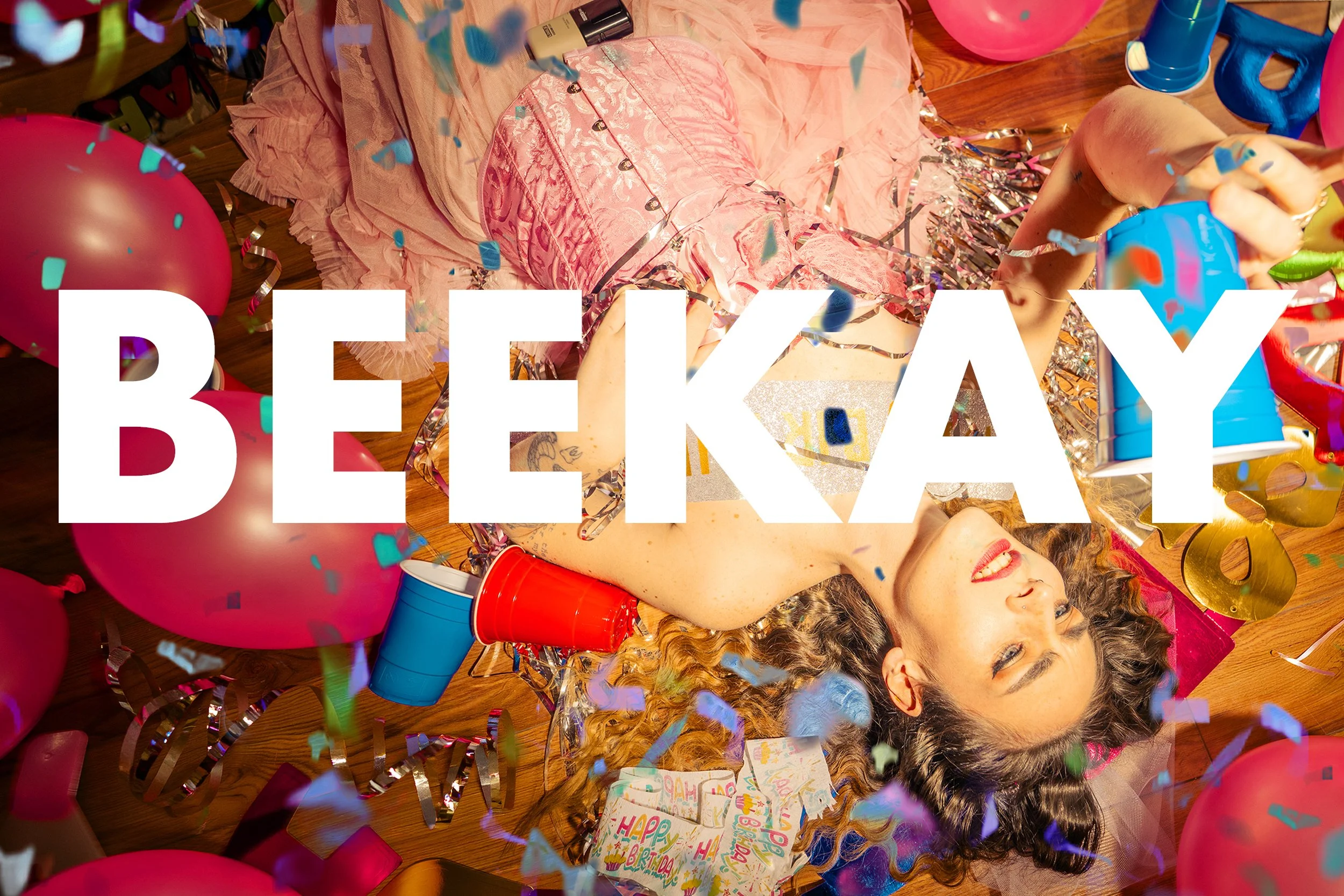 Celebration scene with a young woman lying on the floor, surrounded by balloons, confetti, presents, and birthday decorations.