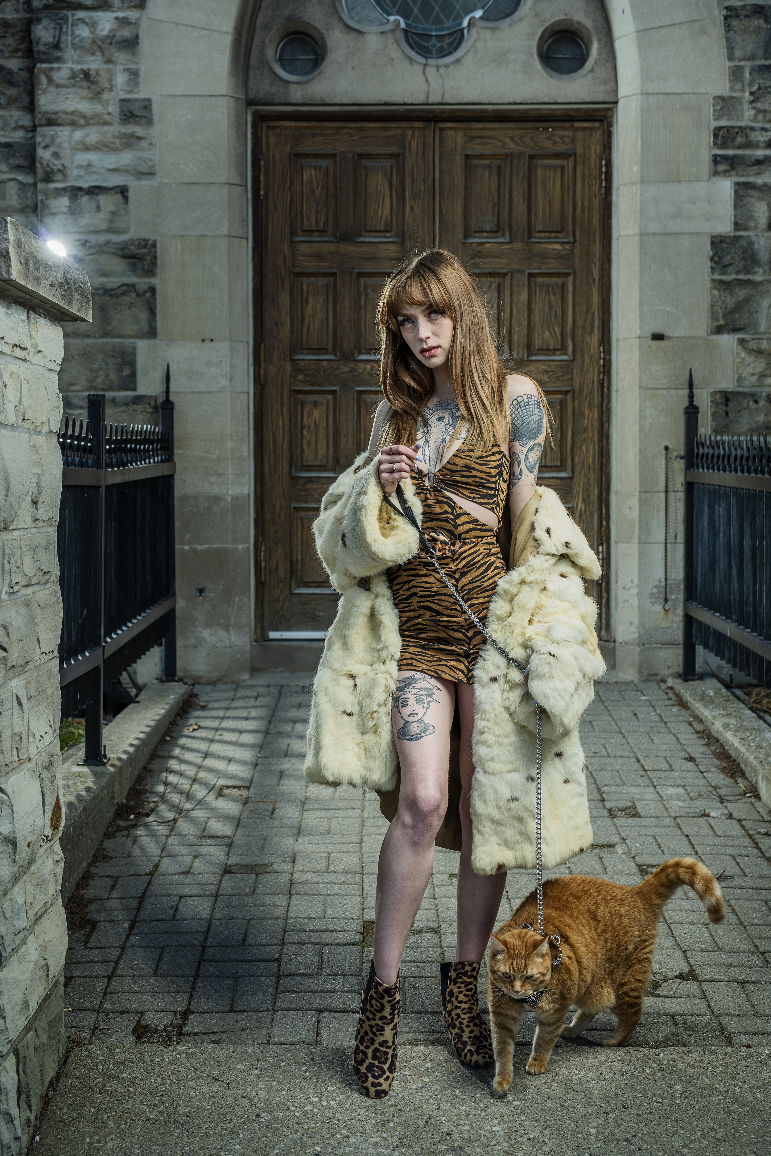 A young woman with long red hair and tattoos, wearing a tiger print dress, a fur coat, and leopard print boots, stands on a sidewalk holding a leash attached to a ginger cat in front of a wooden door.