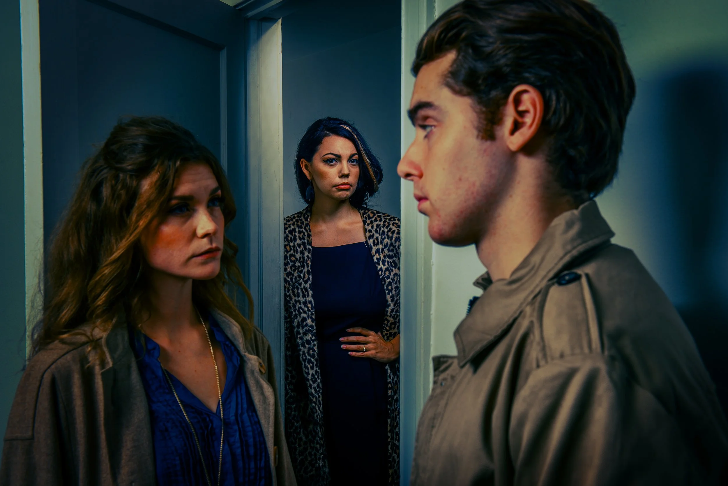 Two women and a man face each other in a small space, with a third woman standing in the background, observing them.
