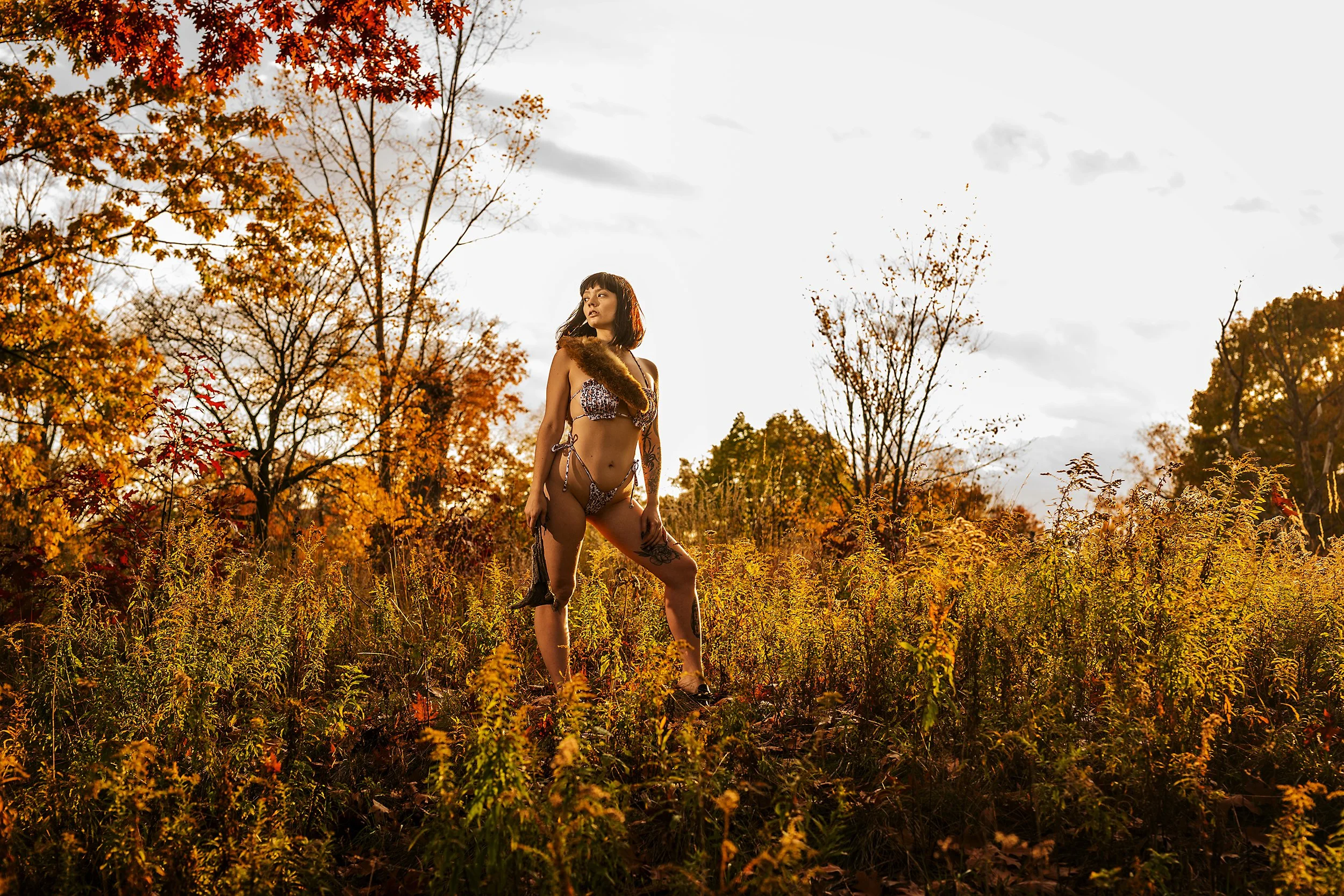 A woman in animal-print bikini with fur scarf posing in a field of autumn foliage during sunset.
