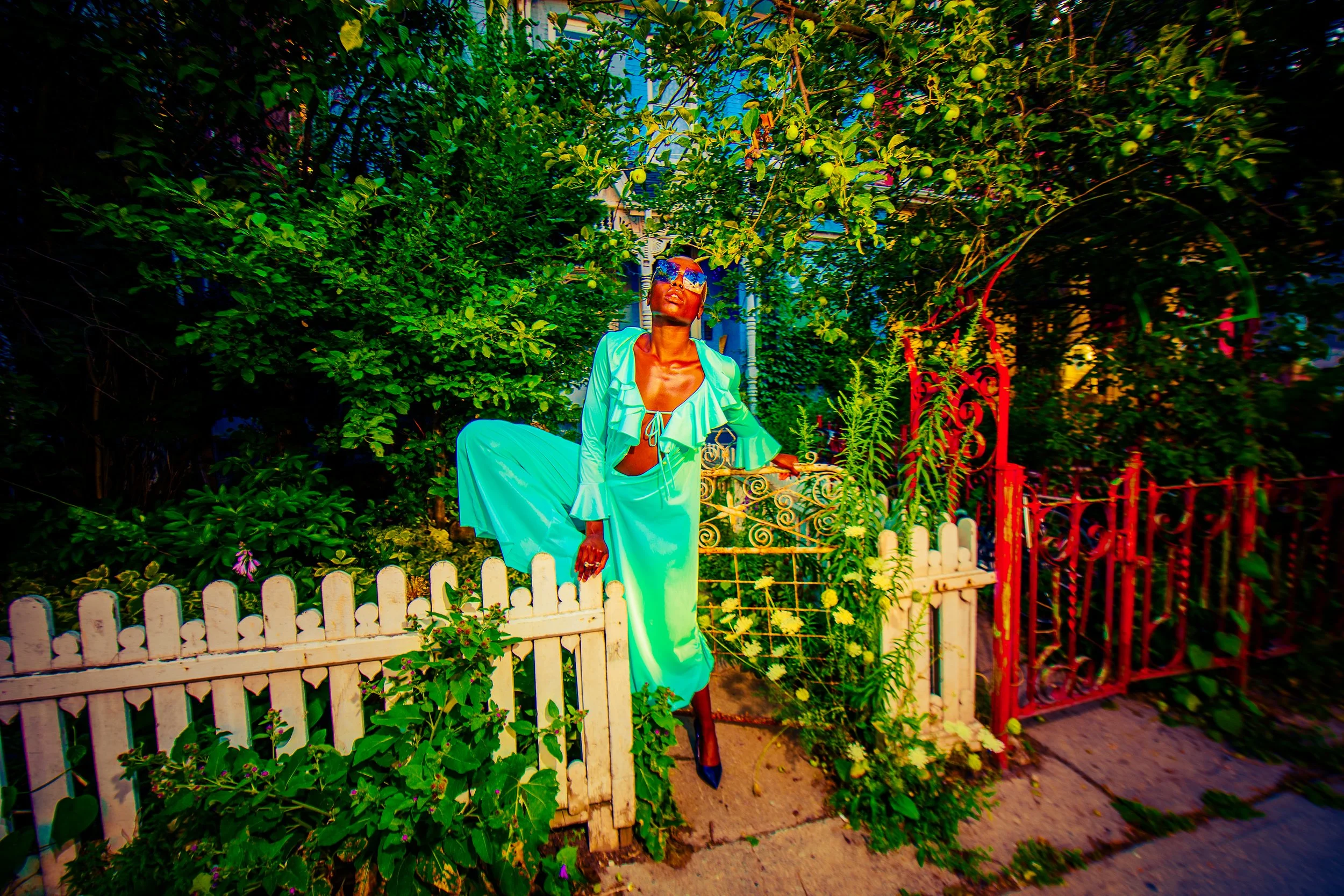 A woman with sunglasses and a teal outfit standing behind a white picket fence in a garden with lush green trees and colorful lighting.