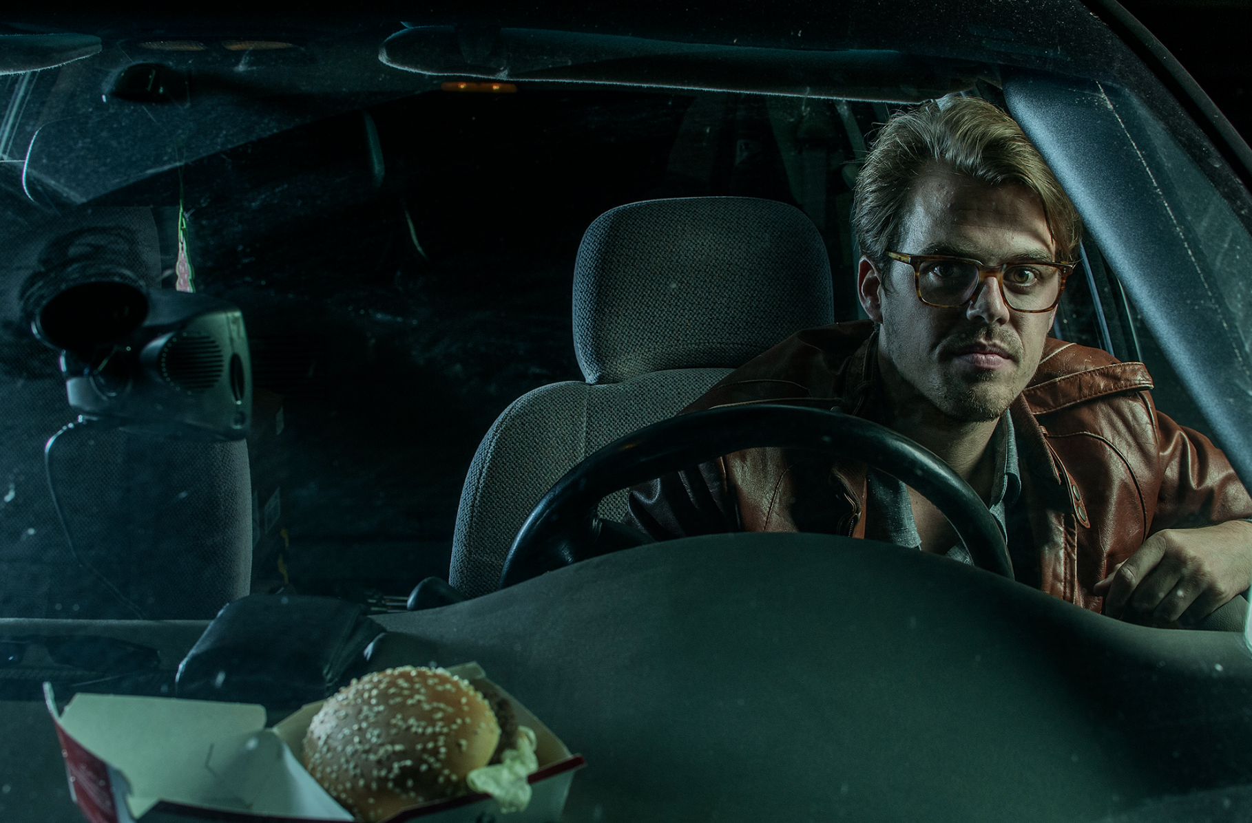 A man with glasses and a brown leather jacket sitting in the driver's seat of a car, looking forward, with a burger in a cardboard container on the dashboard.