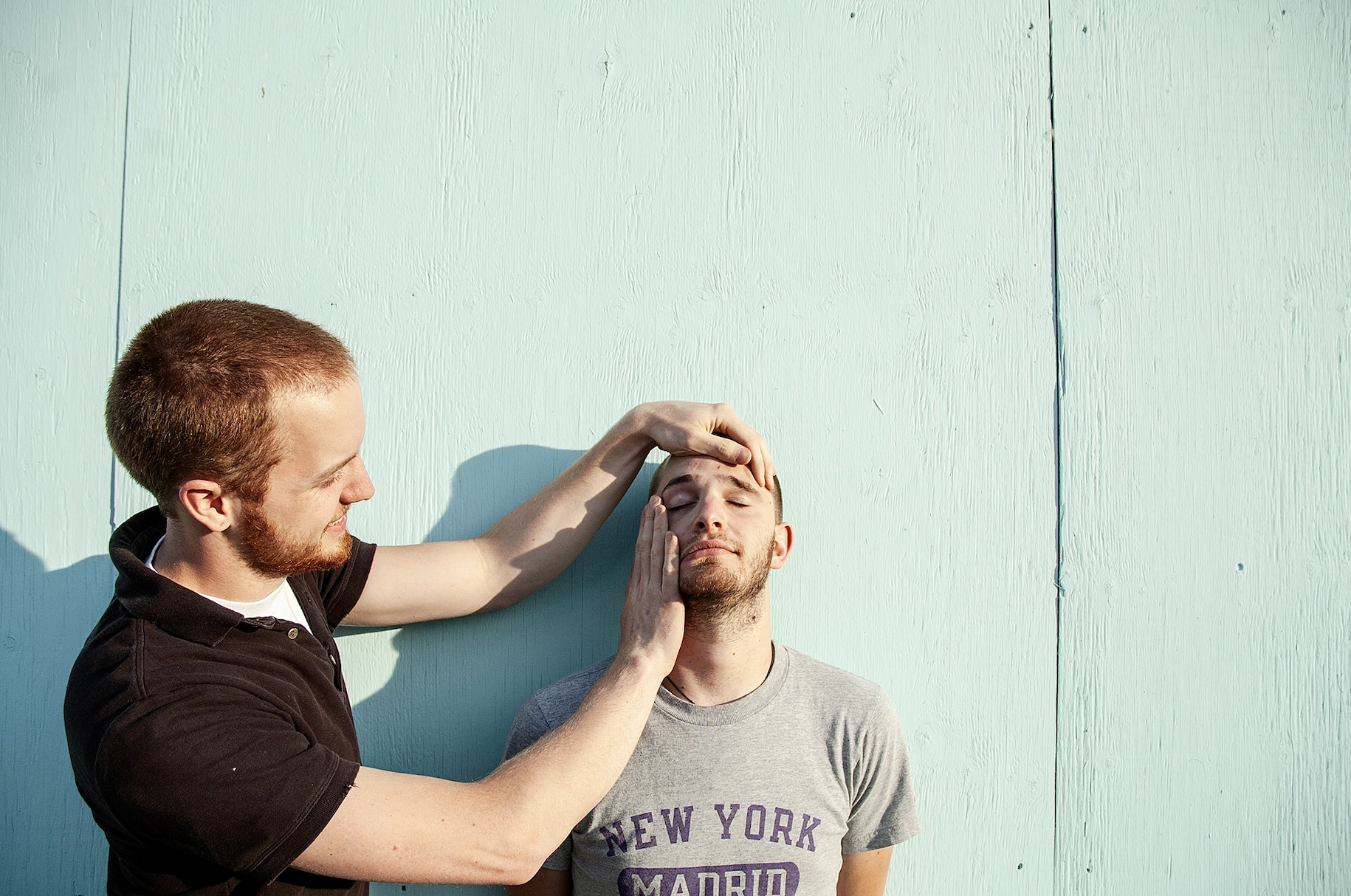 A man with short red hair and a beard is gently touching the forehead of another man with light brown hair and a beard, who has his eyes closed and hand resting on his face, against a light turquoise wooden wall.
