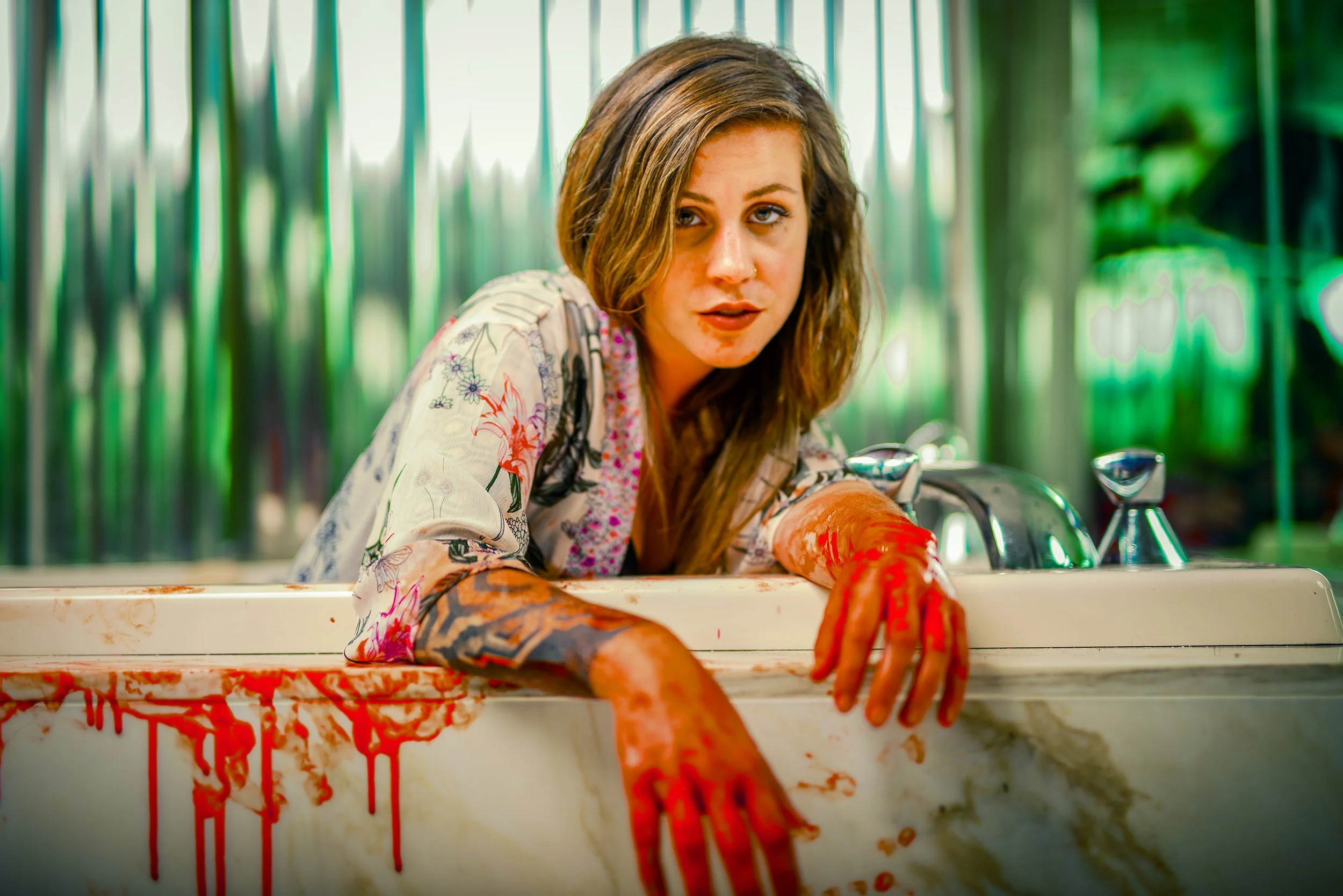 A woman with painted arms leaning over a blood-stained sink, wearing a floral shirt and a serious expression. The background features green vertical bars and foliage.