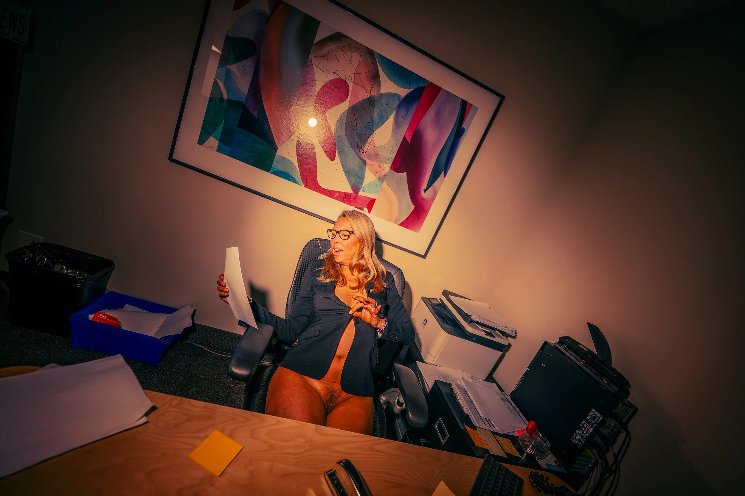 A woman in a black blazer, wearing glasses, is smiling and holding a piece of paper in an office. The office desk has papers, a phone, and office supplies. The background features an abstract, colorful painting on the wall.