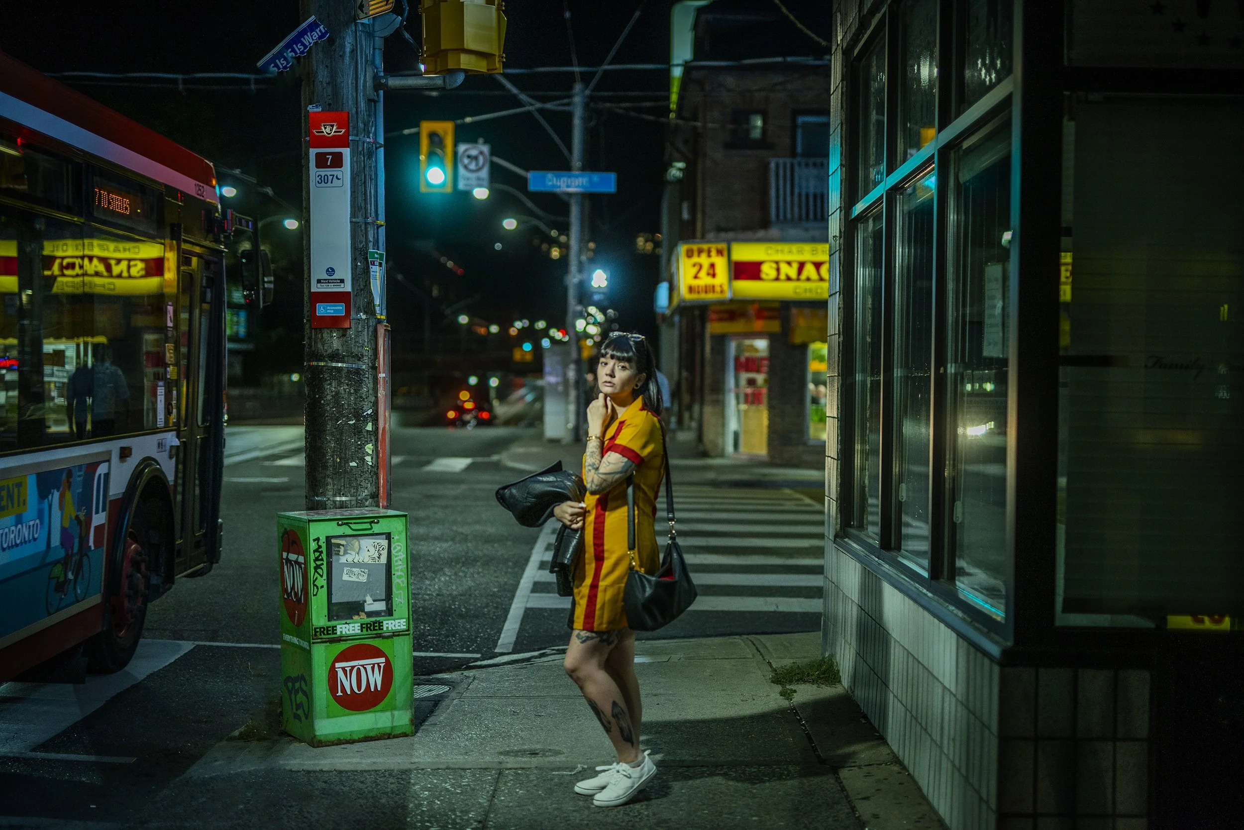 A young woman with tattoos and a ponytail in a yellow and red uniform dress stands on a city sidewalk at night, holding a black bag, with a bus and a corner store in the background, under streetlights and traffic signals.