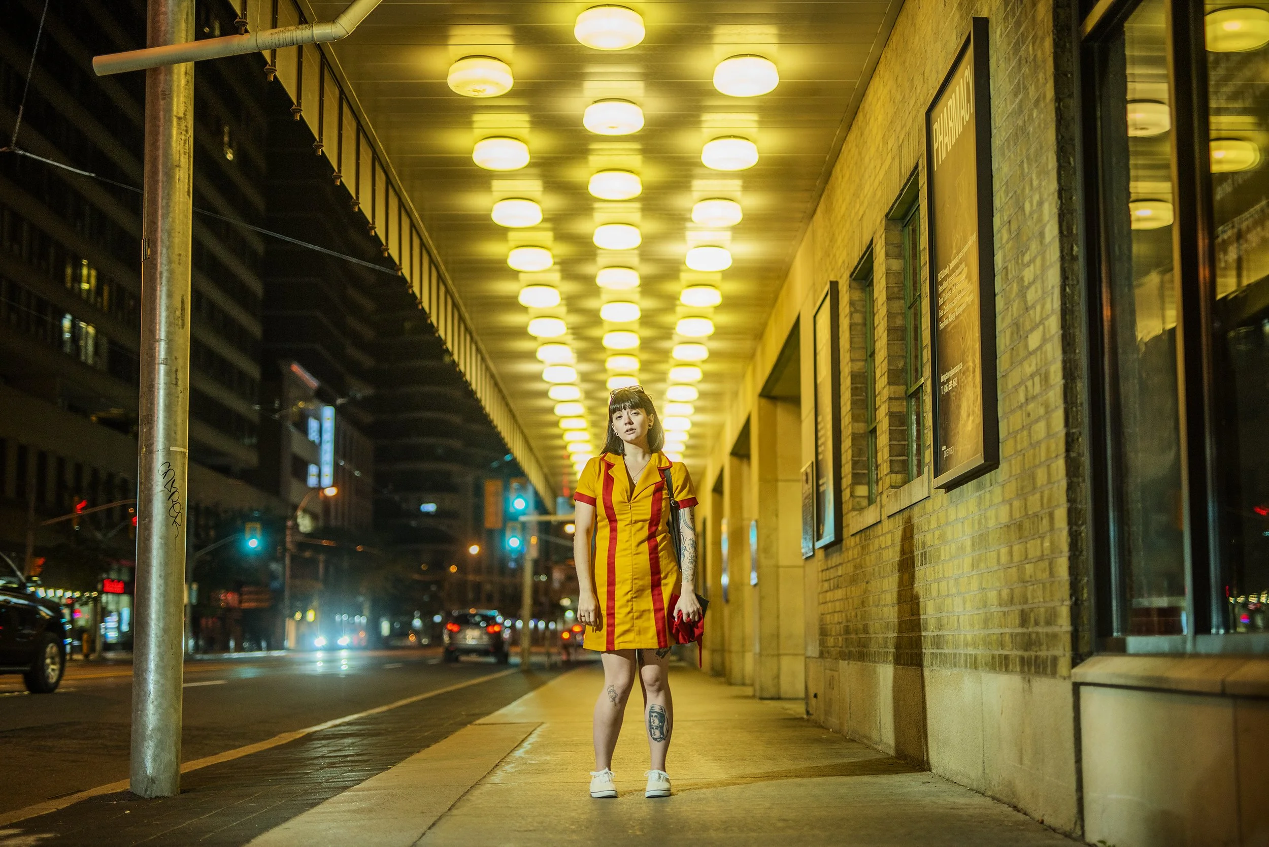 A young woman standing on a city sidewalk at night under bright yellow ceiling lights, wearing a yellow and red striped dress, holding a red clutch, with tattoos on her legs and arms, in front of a brick building.