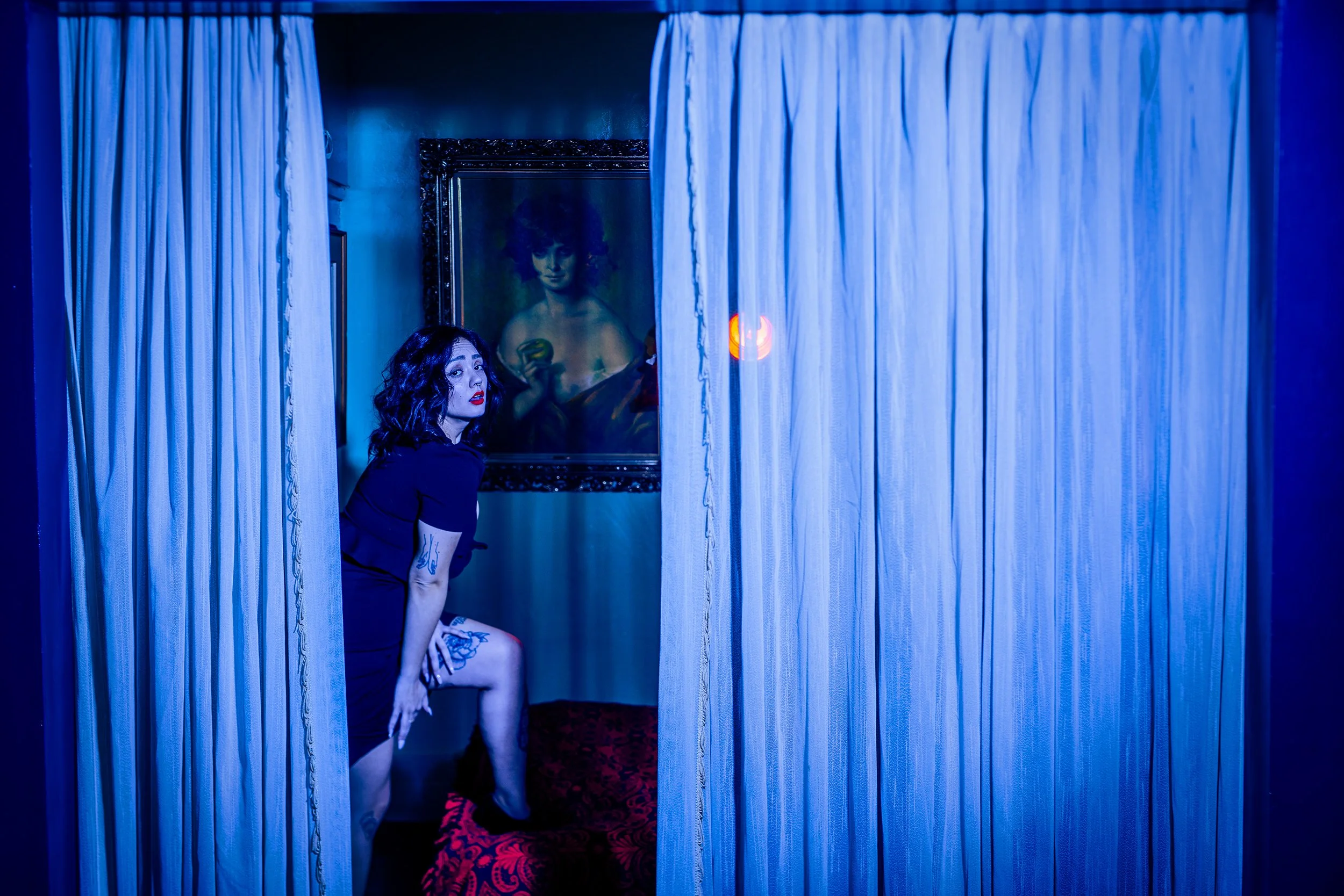 A woman with dark curly hair and red lipstick sits behind a blue curtain, with a framed painting of a woman holding a doughnut visible in the background. The photo has a blue hue.