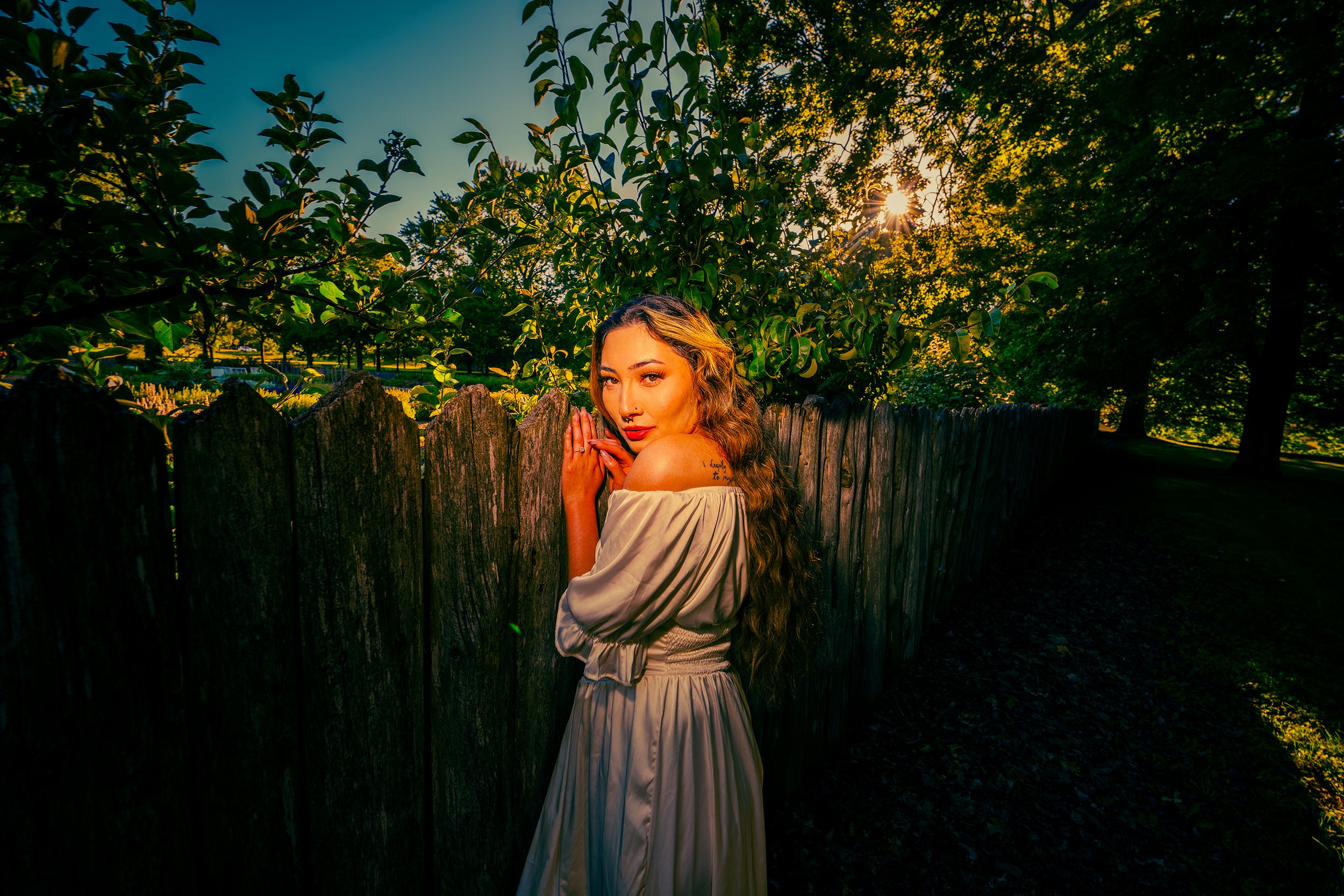 A woman with long curly hair and a light-colored off-the-shoulder dress standing by a wooden fence, with her hands pressed together near her face, in a lush outdoor setting during sunset.