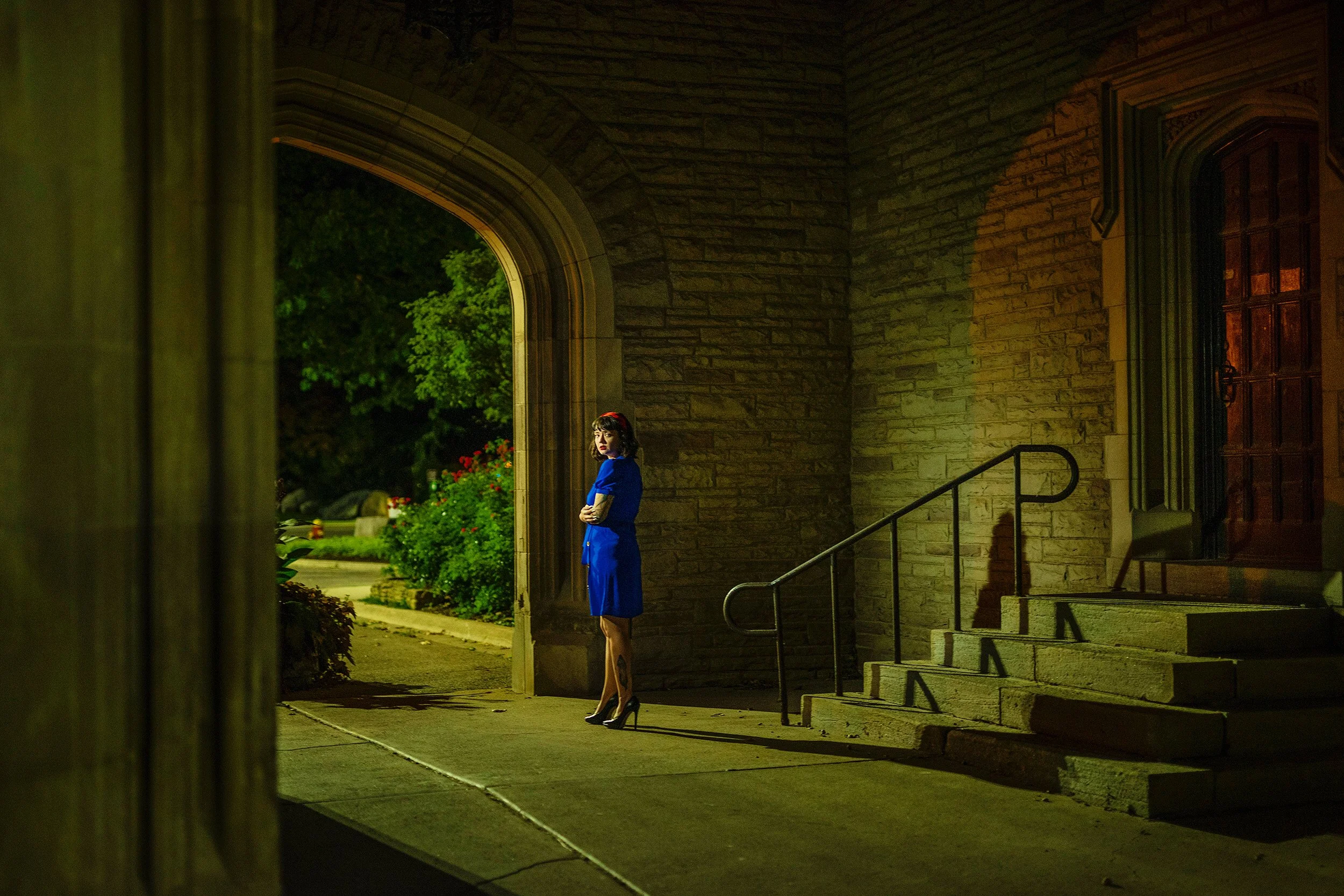 A woman in a blue dress and black high heels stands near the steps of a stone building at night, with green trees and flowers visible outside.