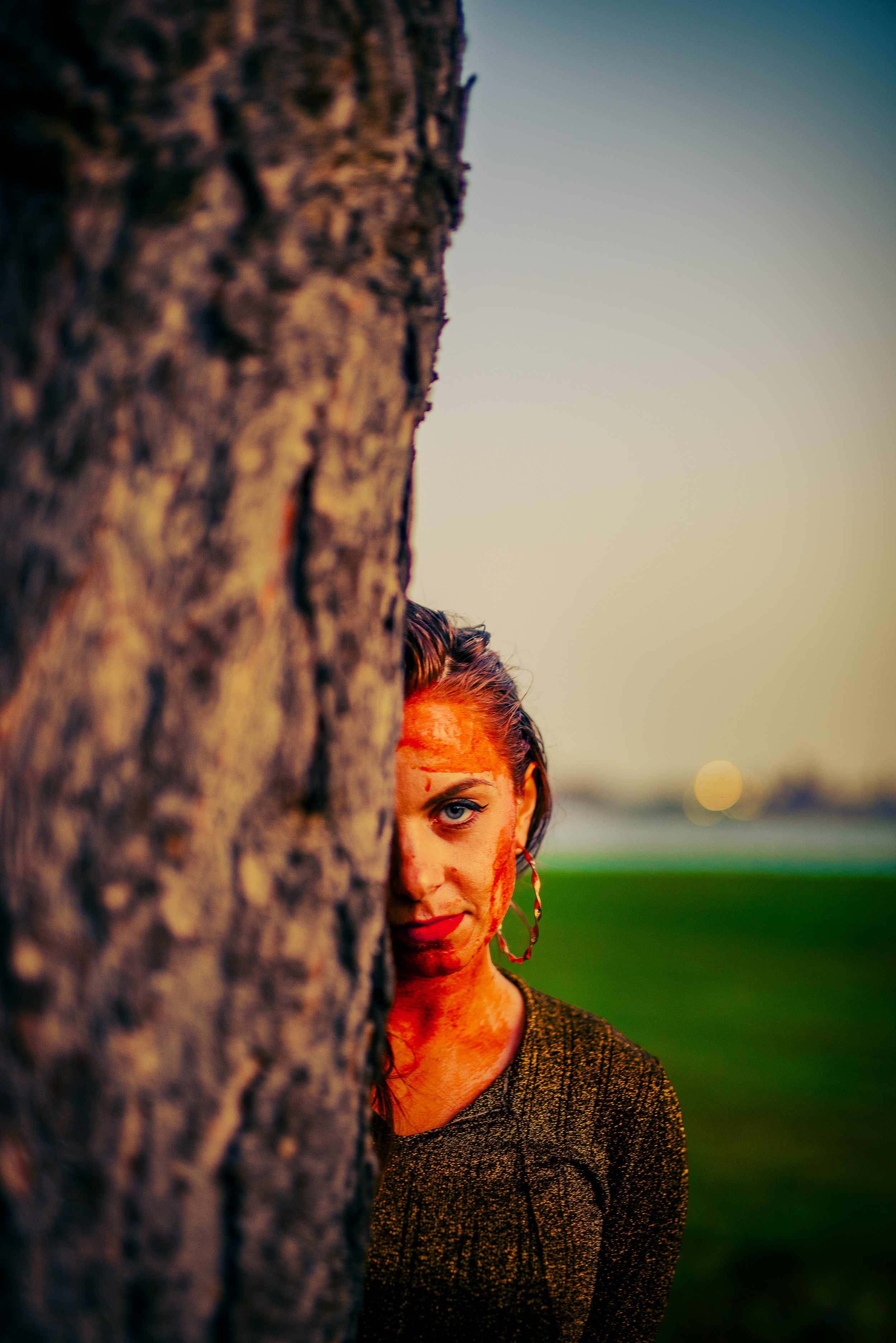 A woman with dark hair and earrings peeking from behind a tree during sunset or dusk, with a blurred landscape and sky in the background.