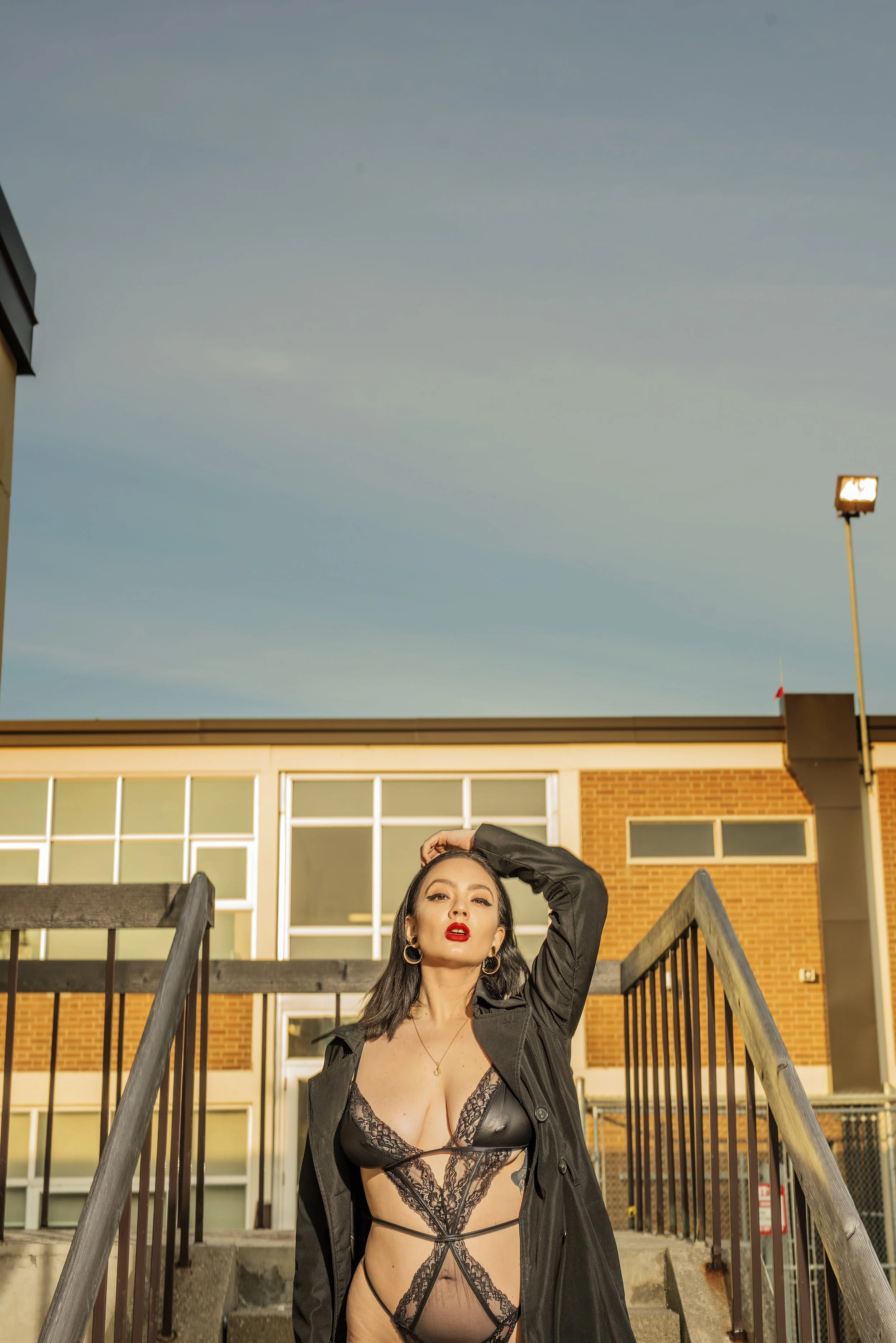 A woman with dark hair, red lipstick, and hoop earrings, wearing black lace lingerie and a black coat, standing on outdoor stairs in front of a brick building during sunset.