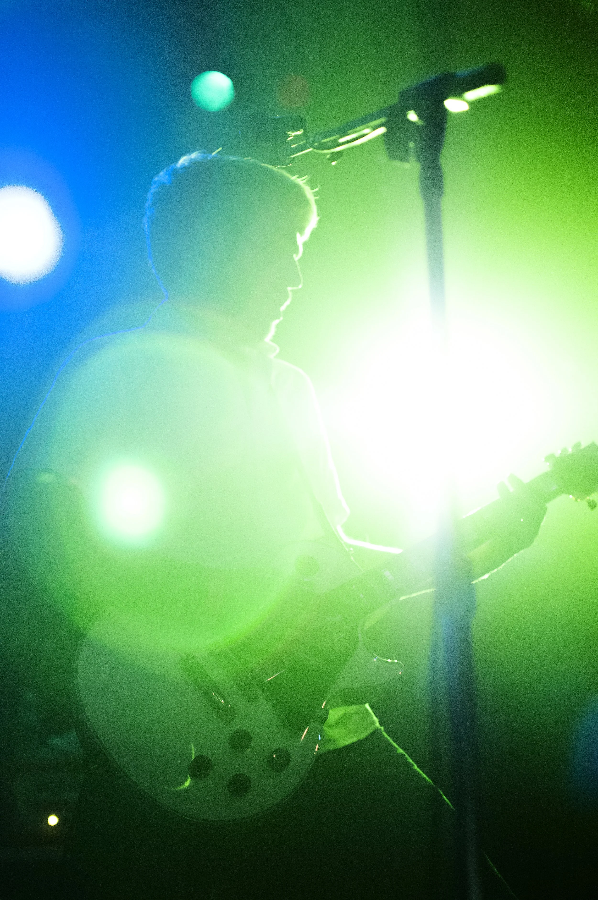 A silhouette of a person playing an electric guitar on stage with colorful green, blue, and white lights creating a hazy atmosphere.