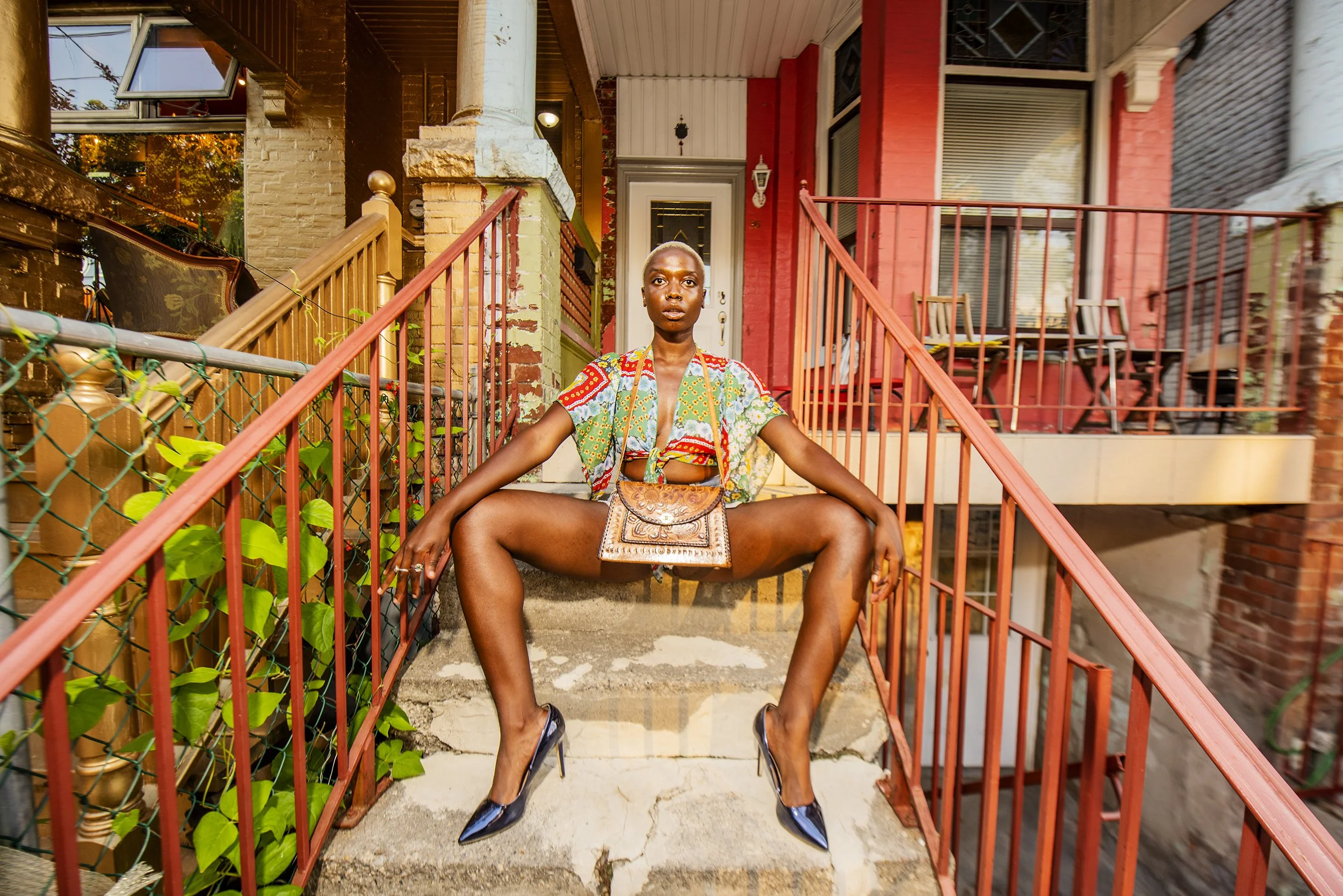 A woman with short hair, wearing a colorful top and black high heels, sitting on stairs with legs apart in front of red houses and a porch, with an intent expression.