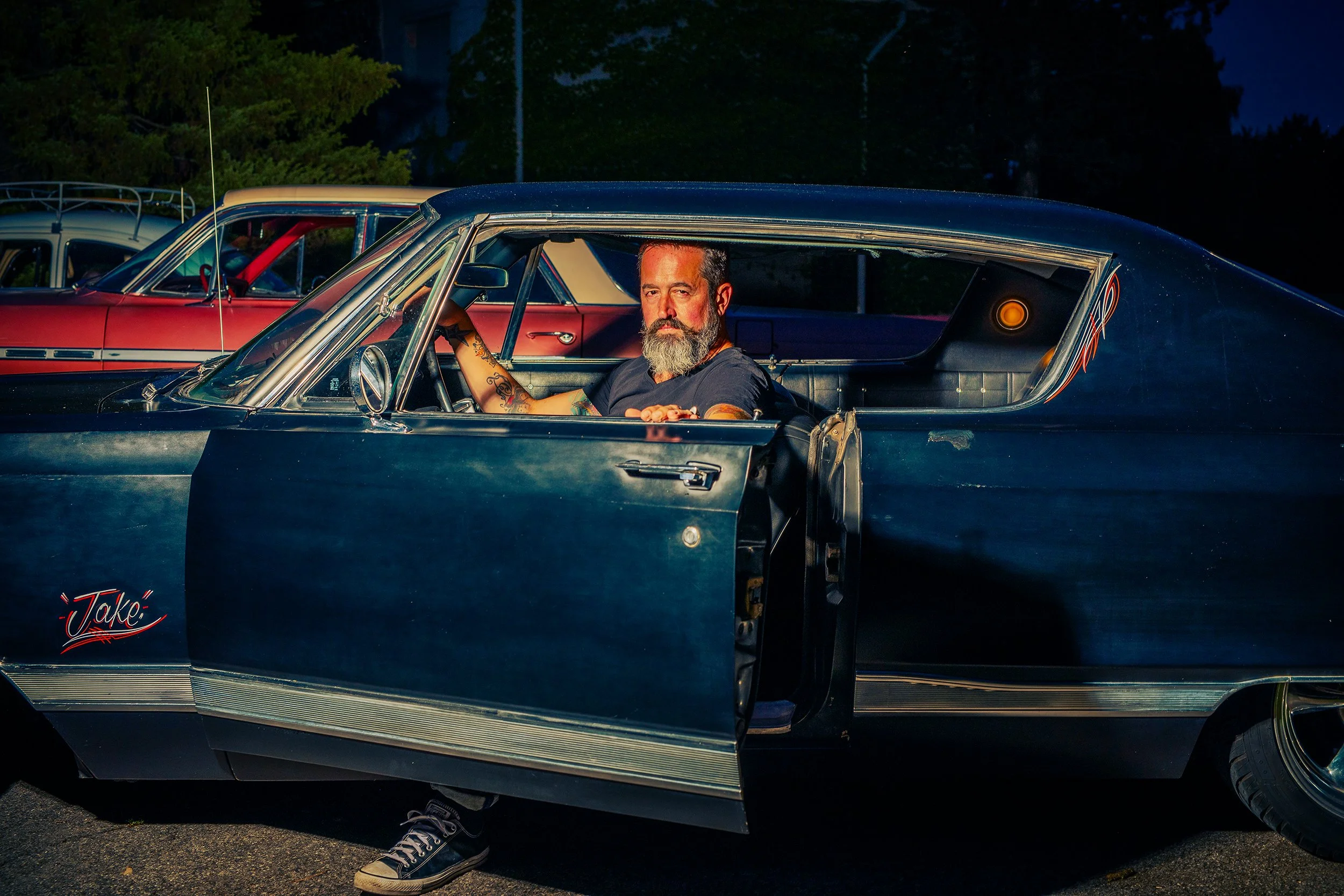 A man with a beard and tattoos sitting inside a vintage black and blue car at night, with the car door open, and other cars parked behind him.
