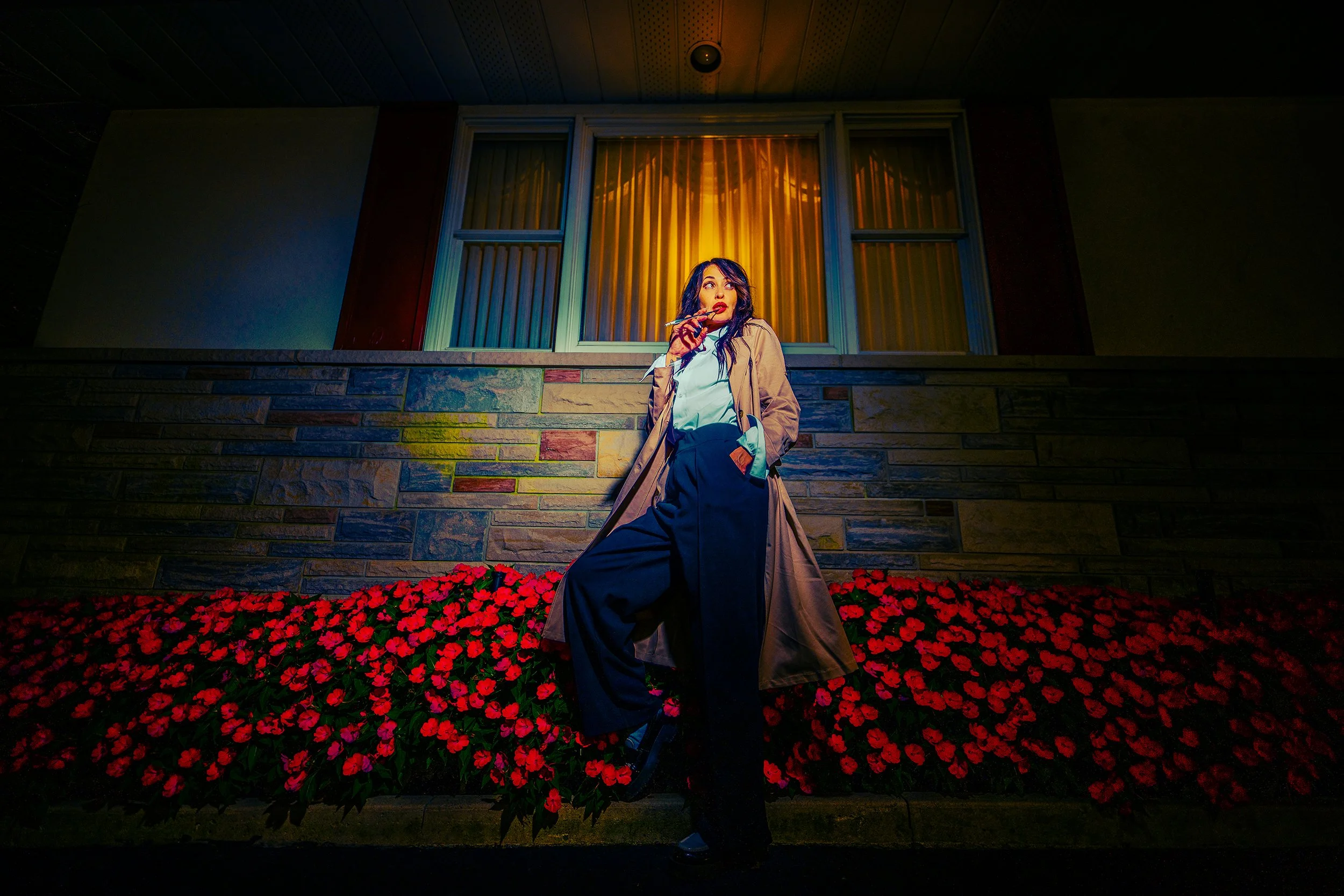 A woman standing in front of a building with a window and stone wall, holding a cigarette, with red flowers at her feet, illuminated by colorful exterior lighting.