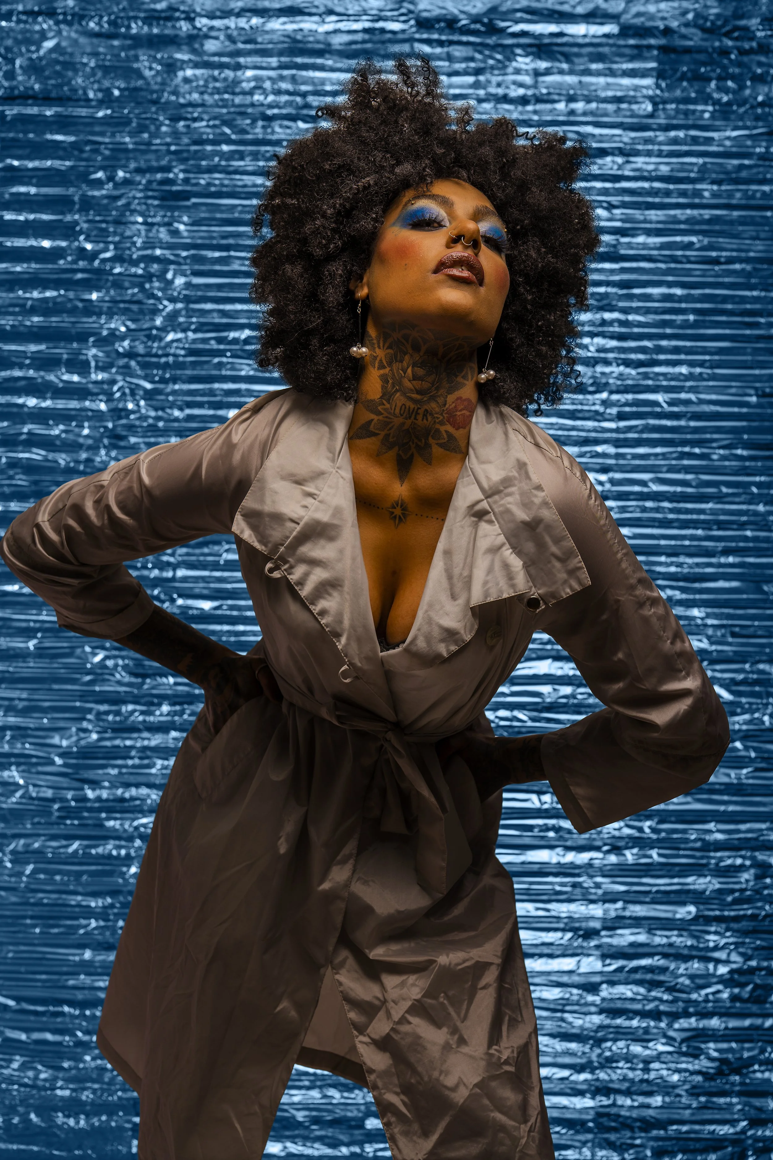 A woman with curly hair, bold makeup, colorful tattoos, and pearl earrings stands in front of a metallic blue textured background, wearing a beige trench coat with her hands on her hips.