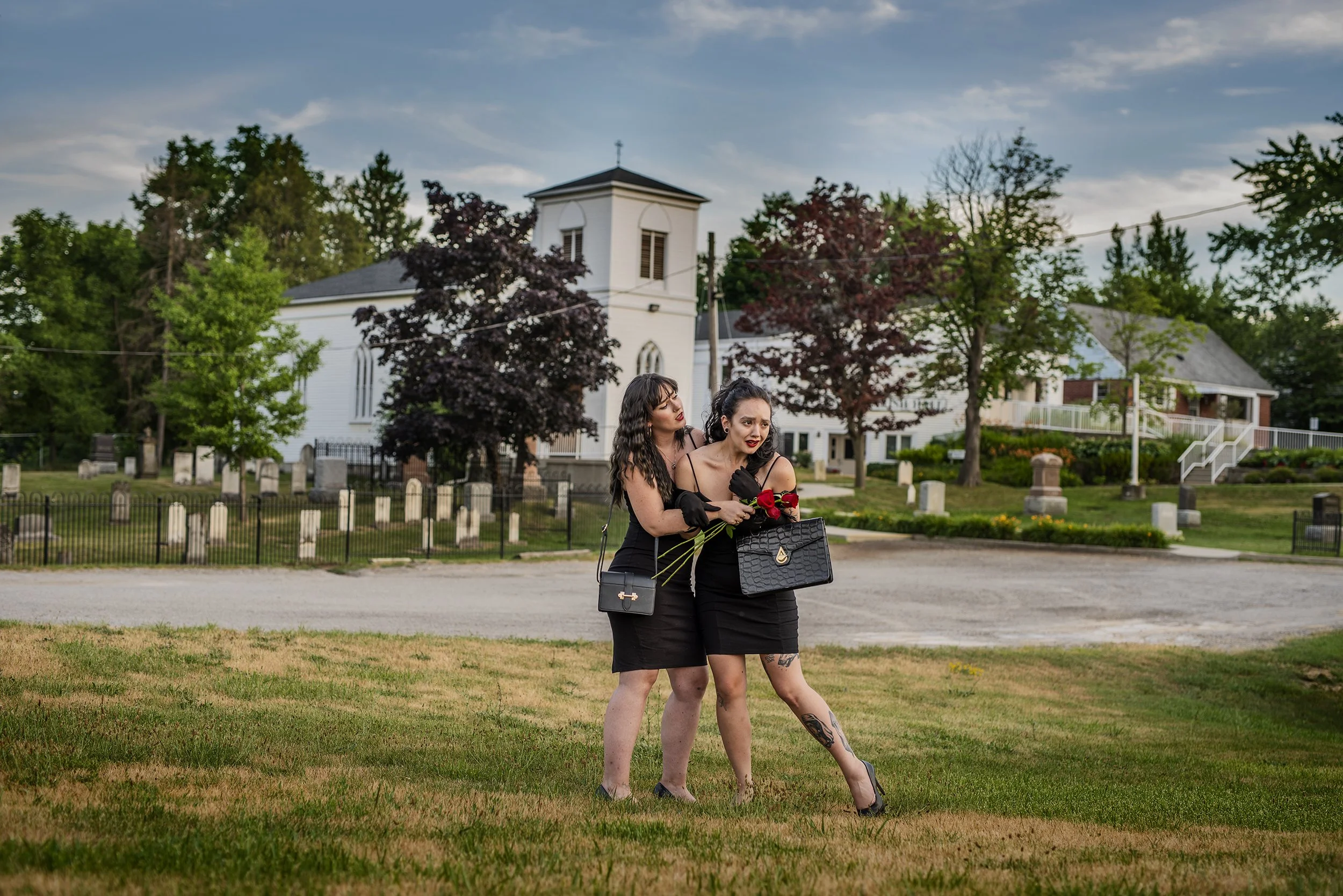 Two women dressed in black, one holding roses and a purse, standing close together outdoors in front of a rural church, with trees and houses in the background.