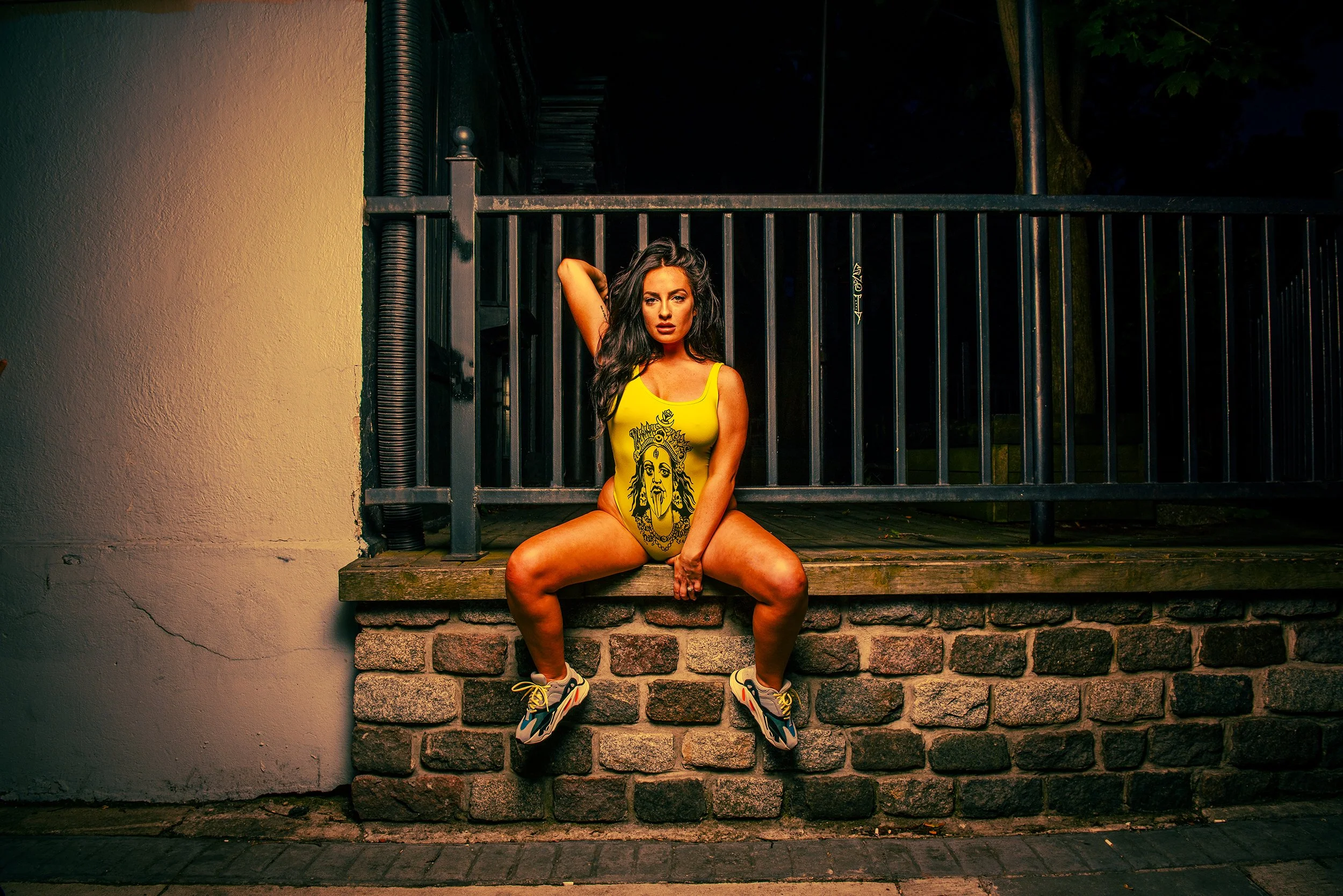 A woman with dark hair sitting on a brick ledge in front of a black metal gate at night. She wears a yellow tank top with a graphic face and athletic shoes, posing with one arm behind her head and the other resting on her knee.