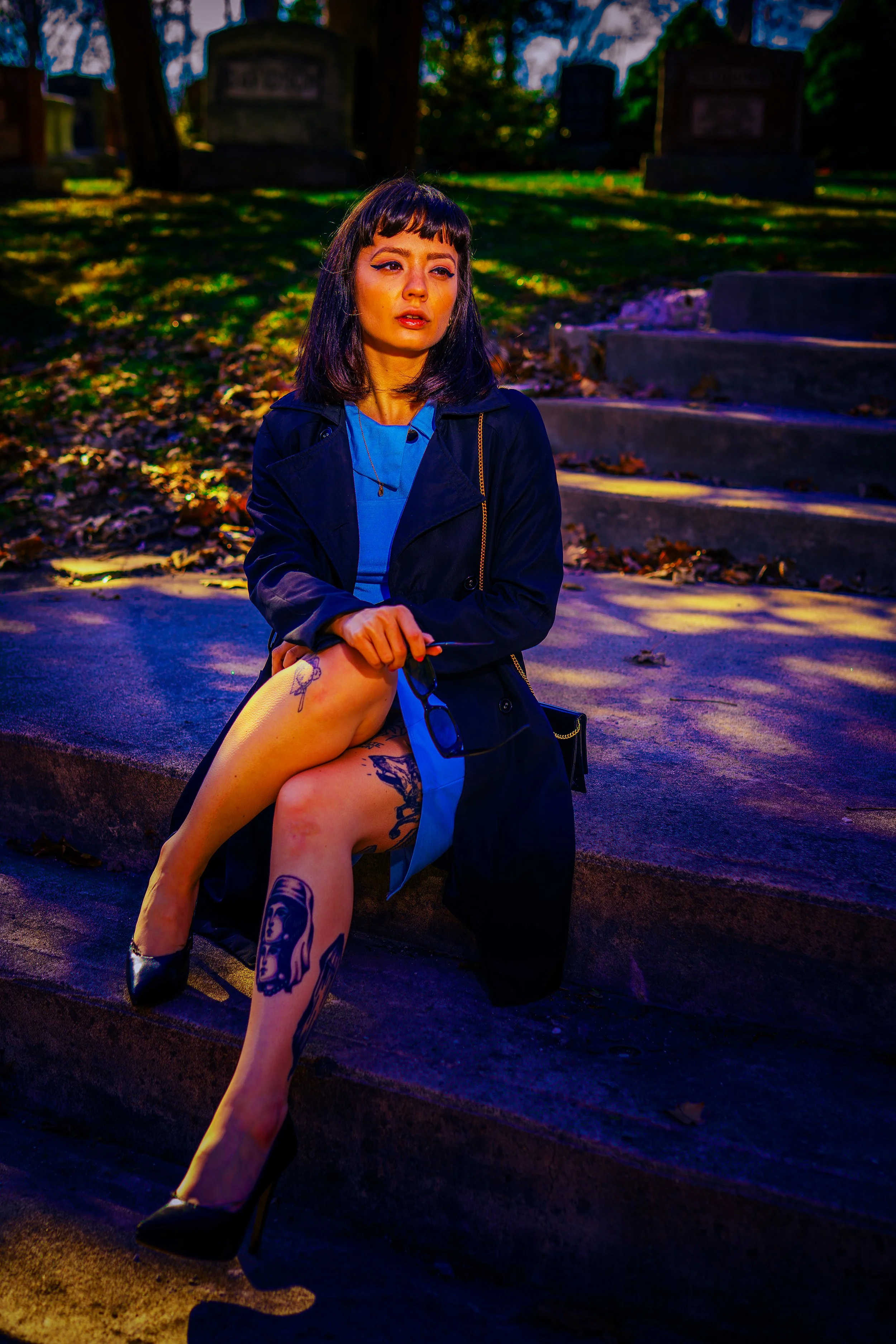 A young woman with dark hair and tattoos on her legs, wearing black heels, sitting outdoors on a concrete surface with autumn leaves and stone steps in the background.