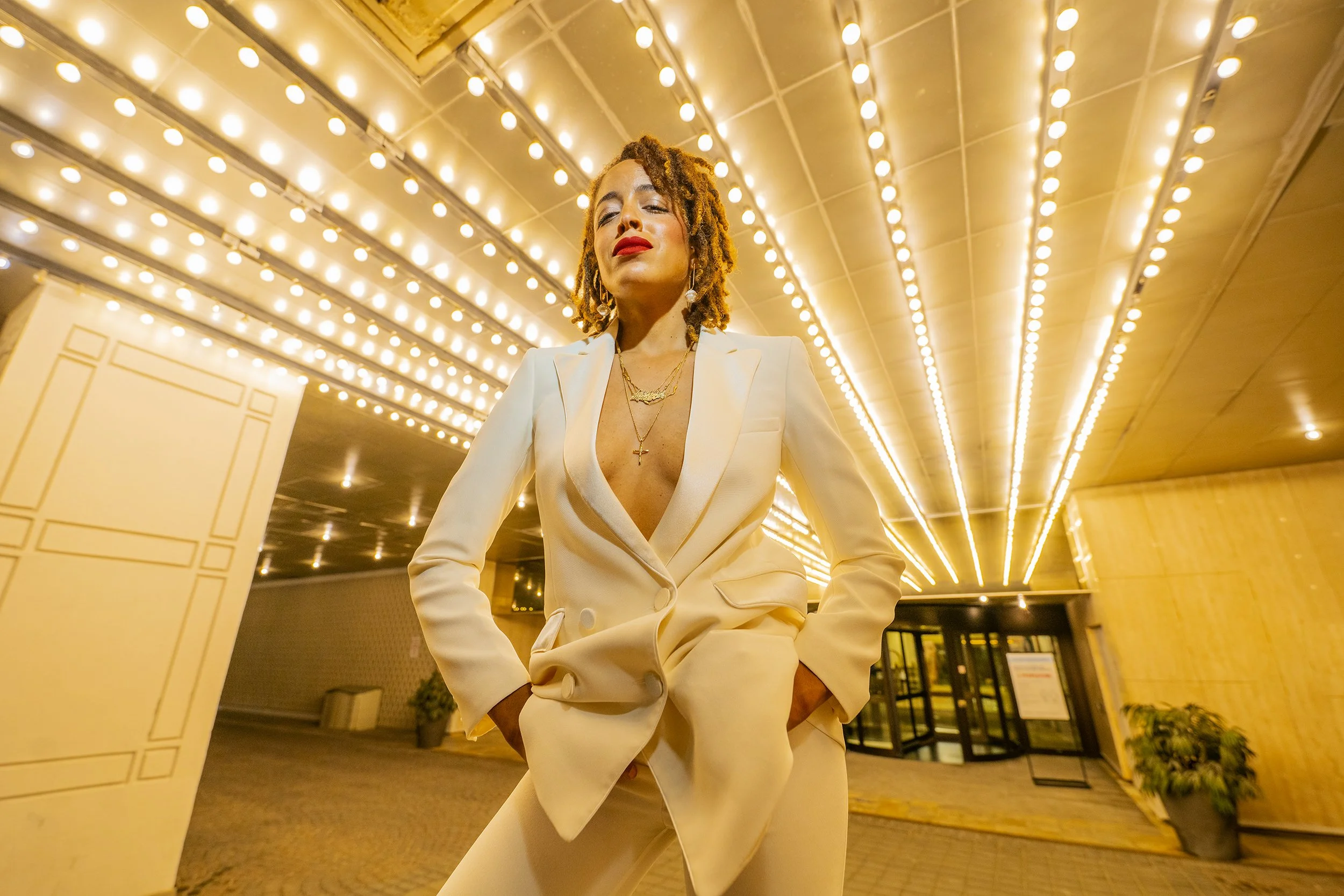 A woman with curly hair and red lipstick wearing a white suit with a deep neckline, standing under a ceiling lined with round white lights in a hotel lobby.