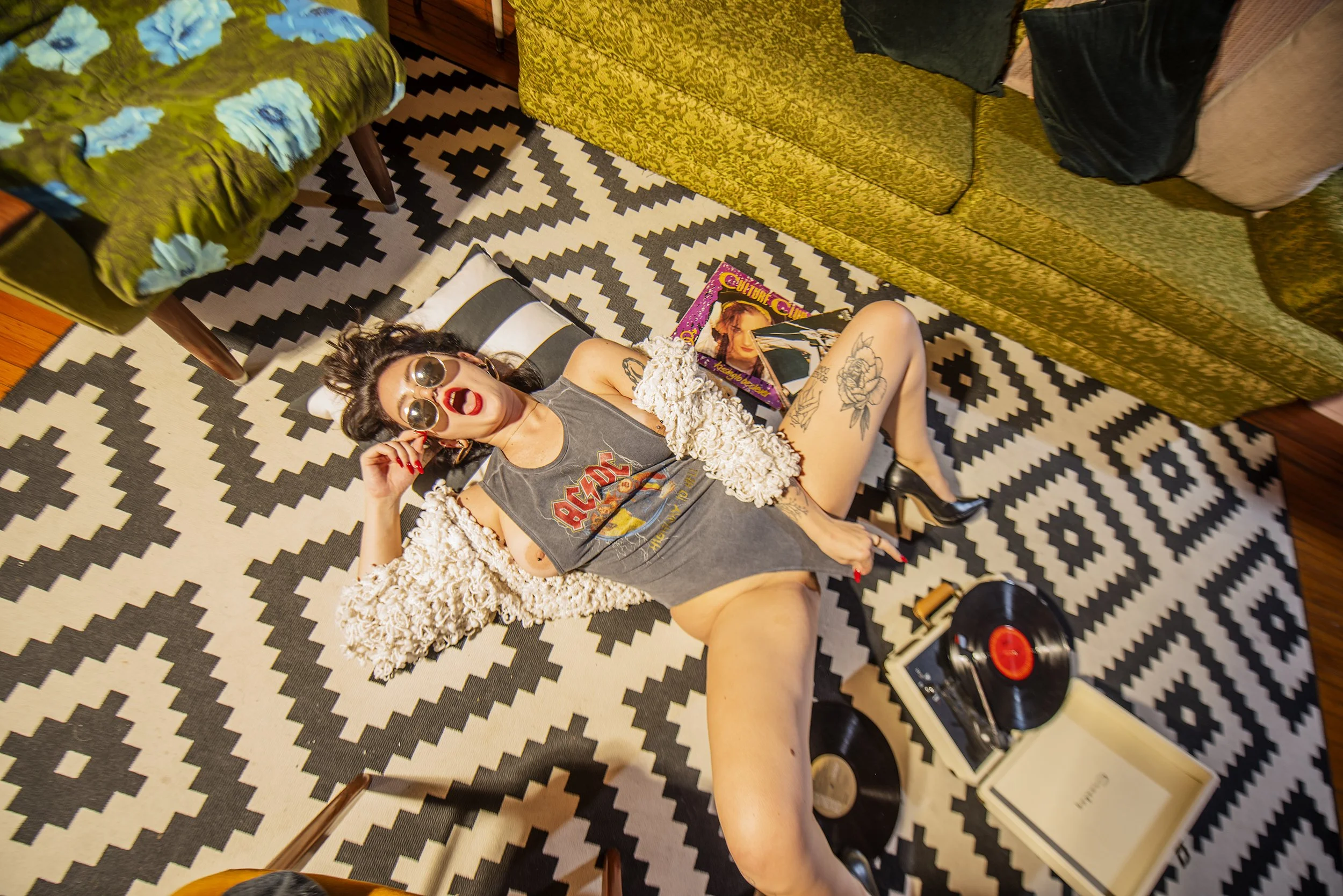 A woman lying on the floor, wearing sunglasses, a ripped T-shirt (AC/DC band logo), and high heels, surrounded by records, a vinyl turntable, a box, and magazines, with a patterned rug and furniture in the background.
