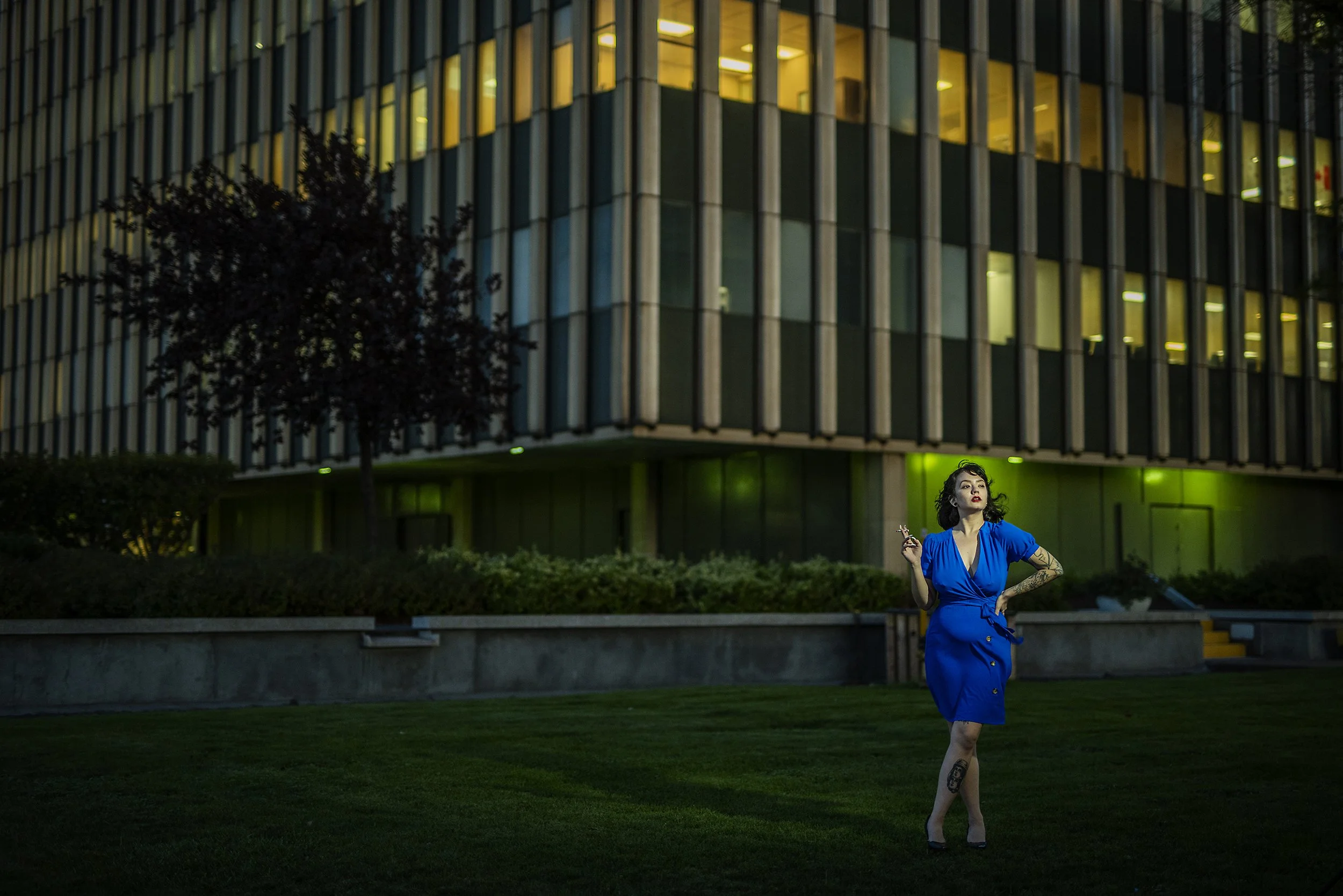 A woman in a blue dress standing on grass outside at night in front of a lit modern office building, holding a cigarette.