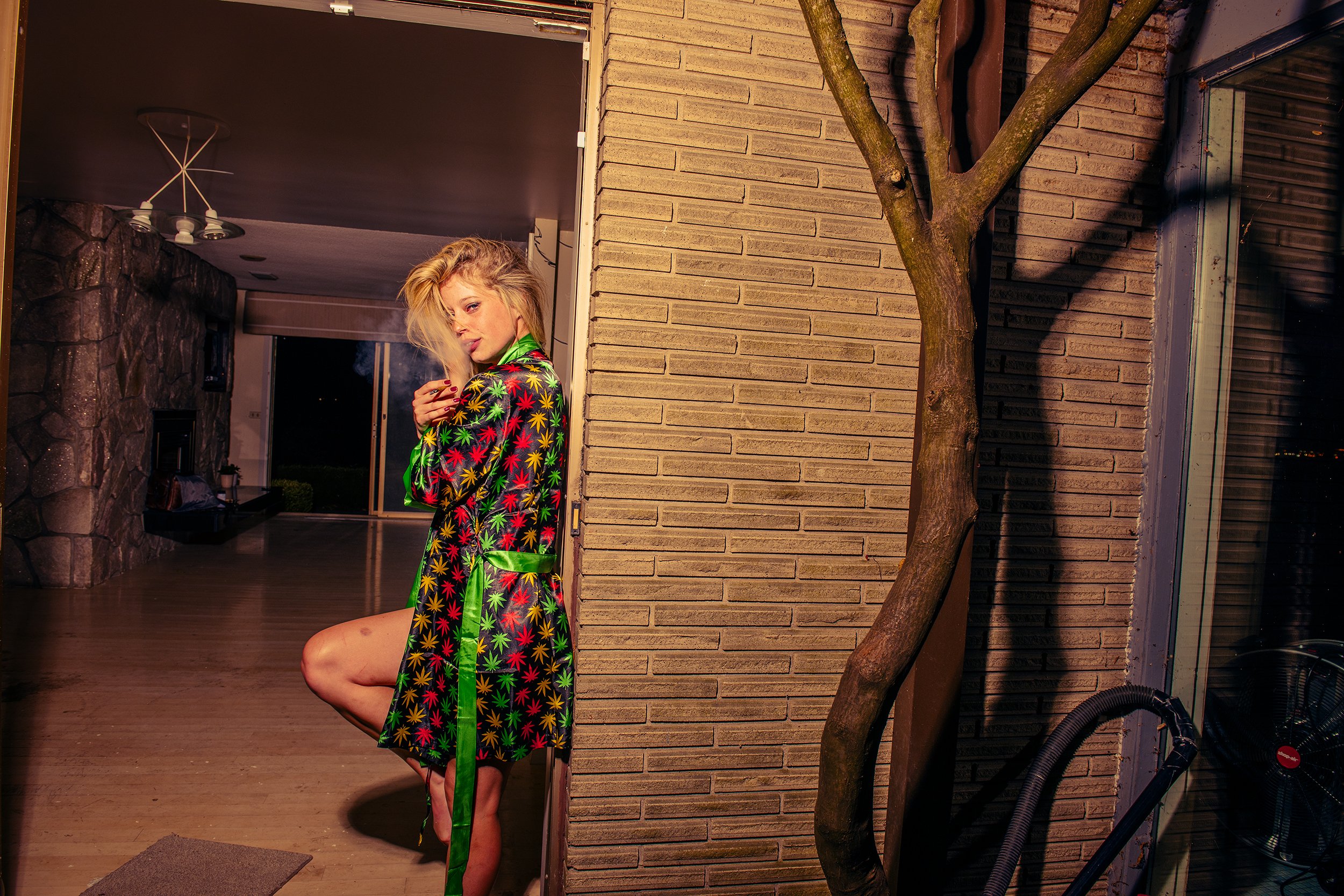 A woman with blonde hair stands next to a brick wall at night, wearing a black robe with colorful marijuana leaf prints and green satin trim, with one leg bent and looking at the camera.