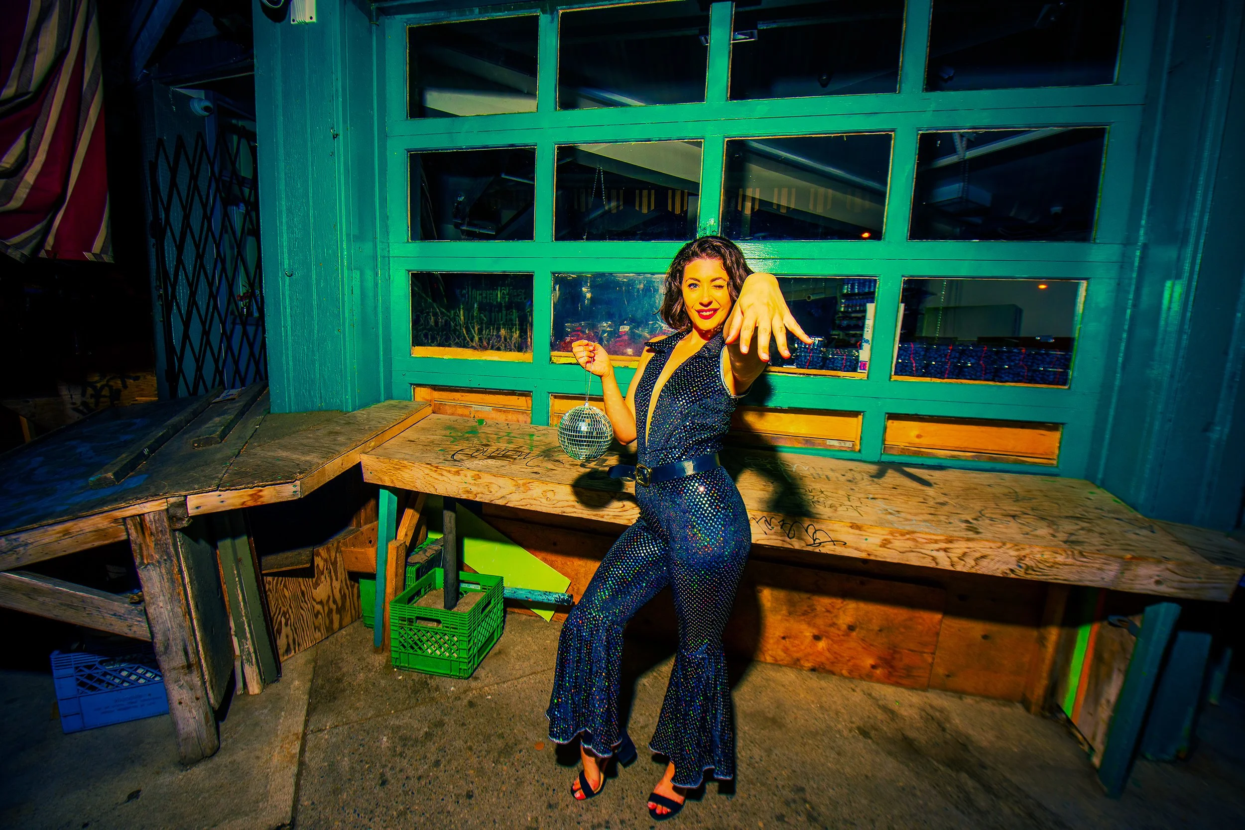 Woman in blue patterned pants and a black top with short wavy hair, smiling and pointing towards the camera, standing in front of a turquoise garage door. There are wooden platforms and green crates around her.