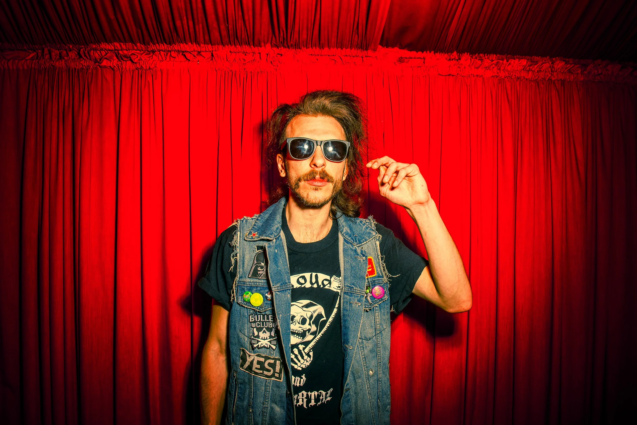 Person with long hair, wearing sunglasses, a black t-shirt with white skull graphic, and a denim vest with colorful pins, standing in front of red curtains.