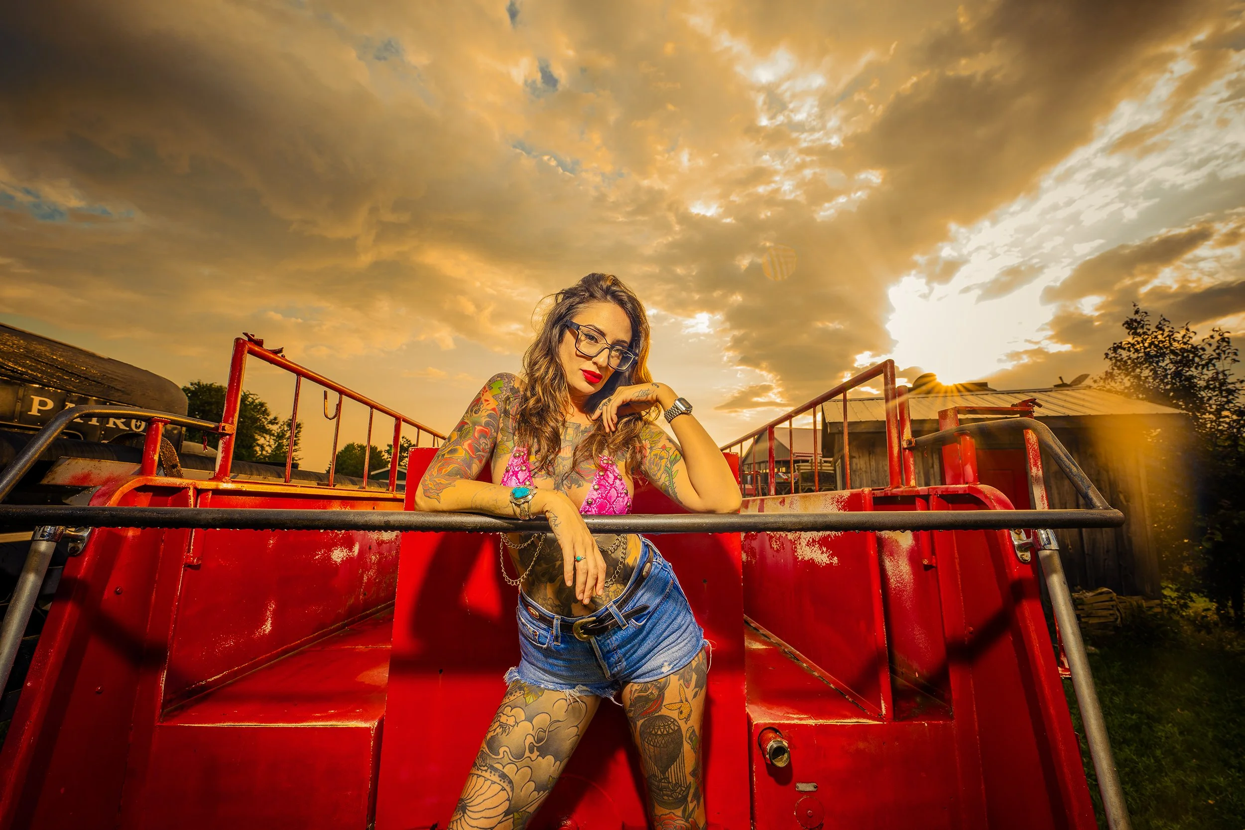 A woman with tattoos, glasses, and red lipstick leaning on a red fire truck at sunset