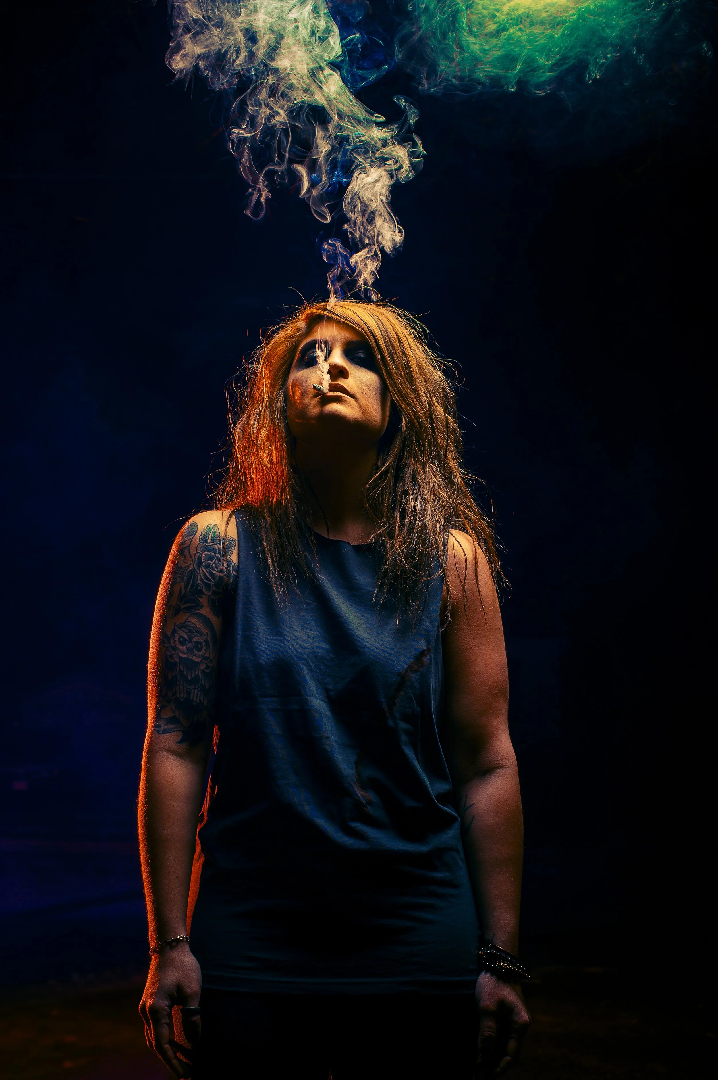 A woman with long orange-brown hair and tattoos on her arm, wearing a sleeveless dark top, standing in a dark setting with colorful lights. Smoke or vapor rises from her mouth, creating a cloud above her head.