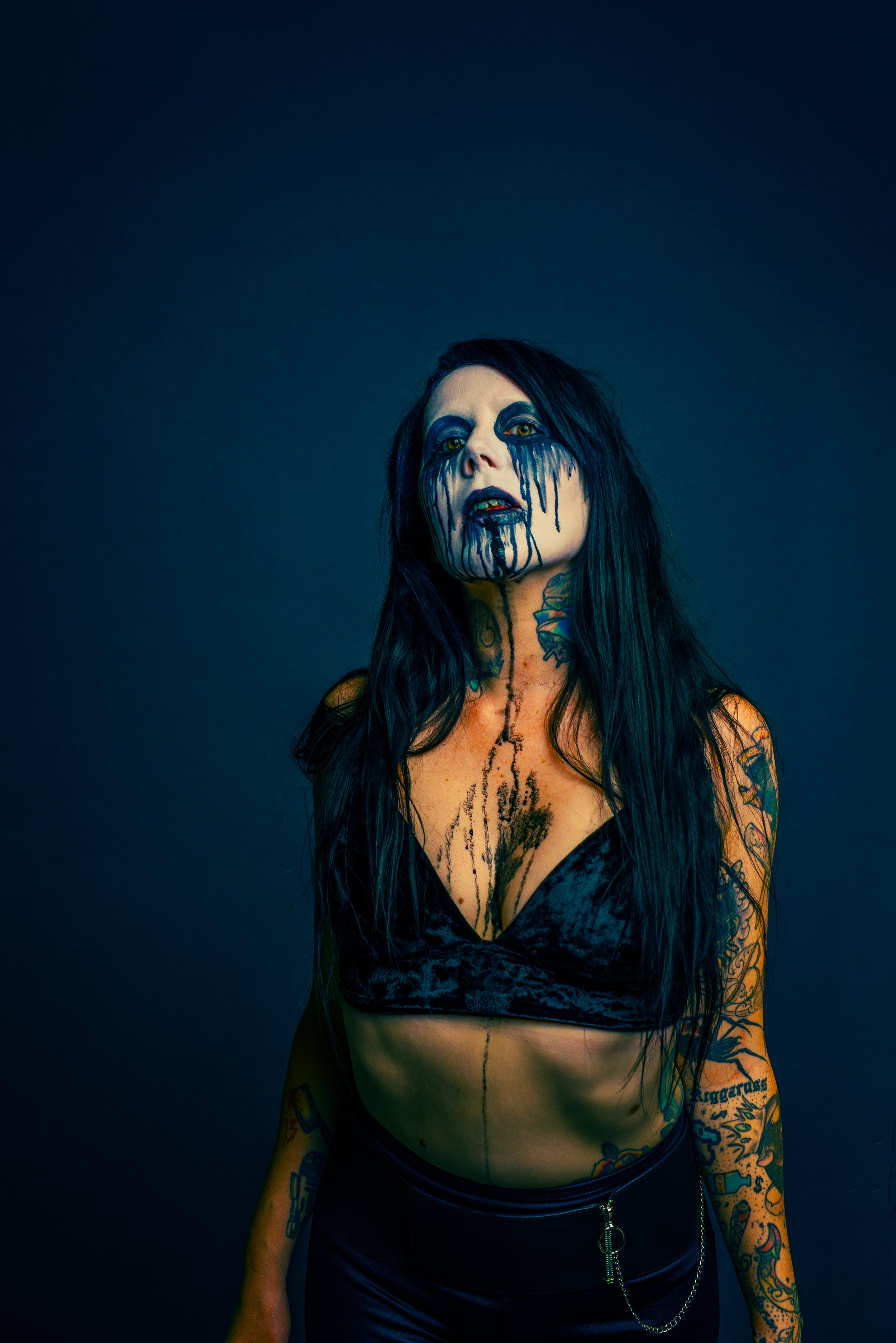 A woman with long black hair and yellow eyes, covered in blue face paint that drips down her face, neck, and chest, wearing a black velvet top and a black leather skirt with a zipper and chain, against a dark background.