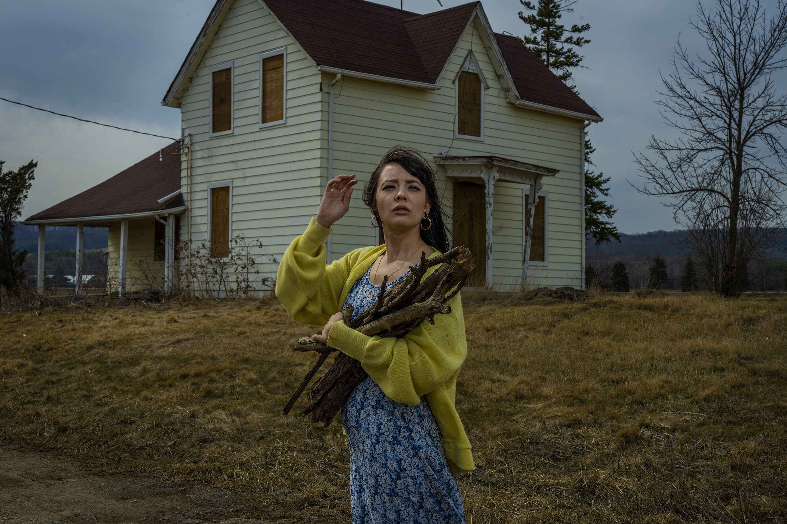 A woman with dark hair wearing a yellow cardigan and blue floral dress holding a bunch of sticks or wood, standing in front of a dilapidated, boarded-up house with peeling paint and missing windows, in a rural area with overcast sky and bare trees.