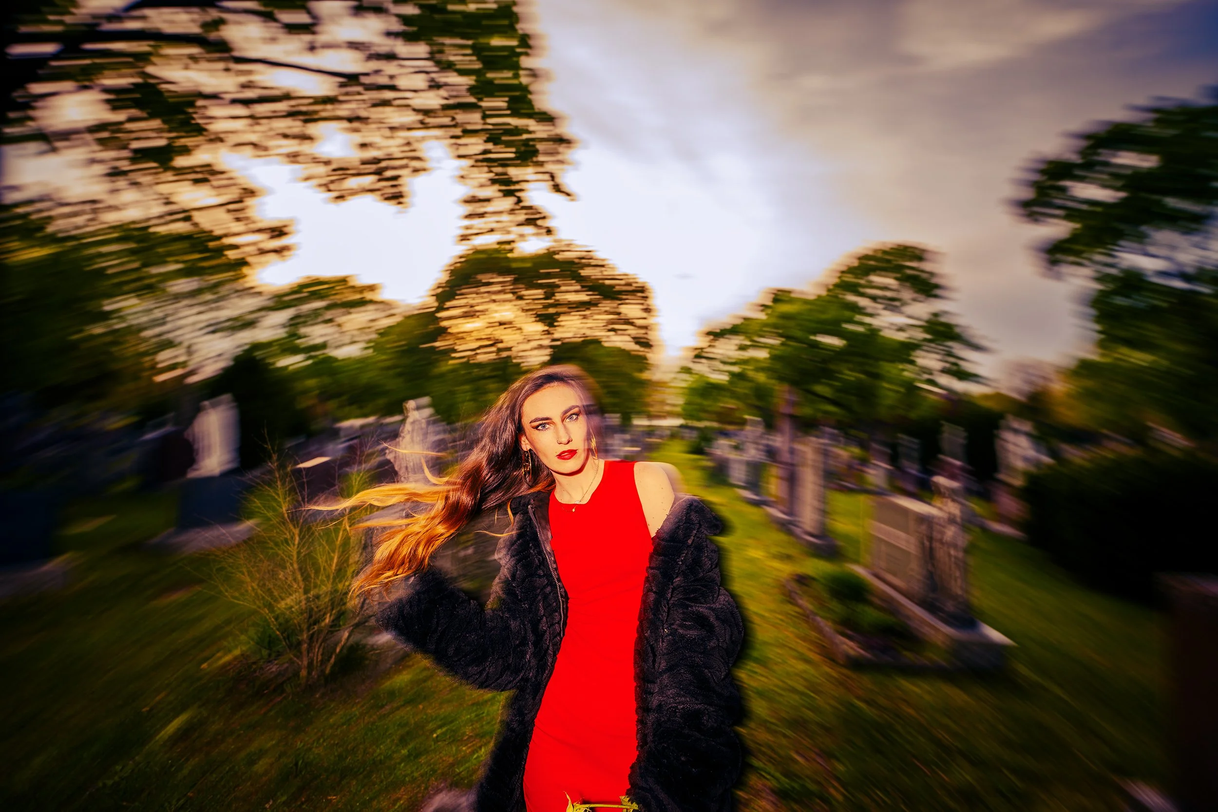 A woman with long flowing hair wearing a red top and black jacket stands outdoors in front of trees during sunset, with a motion blur effect creating a circular swirl around her.