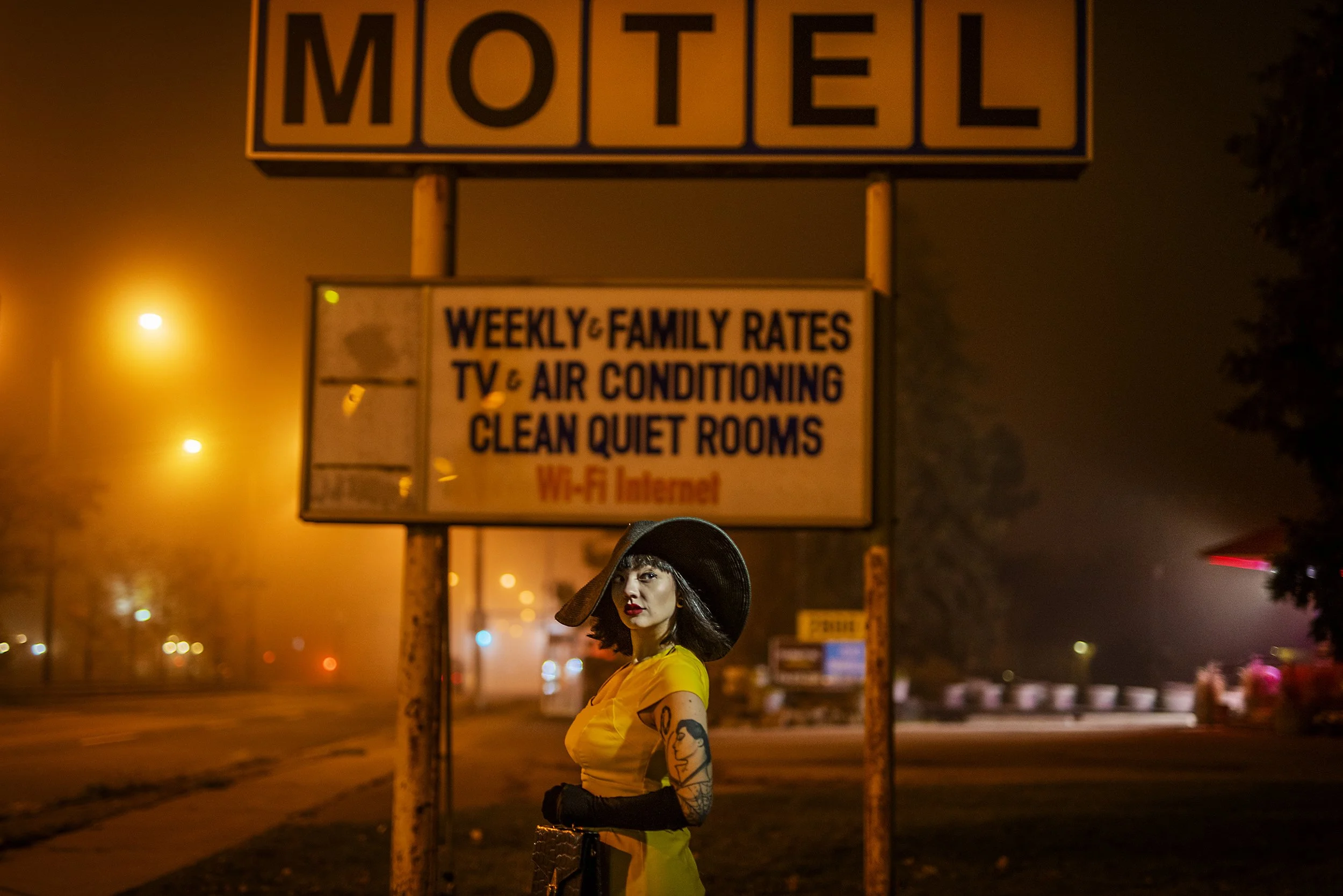A woman with tattoos on her arm wearing a yellow dress, black gloves, and a large black hat stands on a foggy street at night in front of a motel sign. The motel sign reads 'MOTEL' and advertises weekly and family rates, TV, air conditioning, clean q
