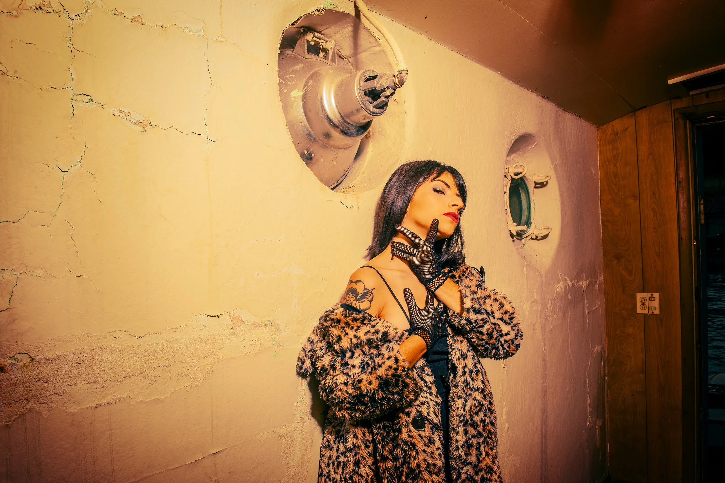 A woman with dark hair and bold makeup posing against a textured yellow wall with cracks, wearing a leopard print fur coat, black gloves, and striking red lipstick.