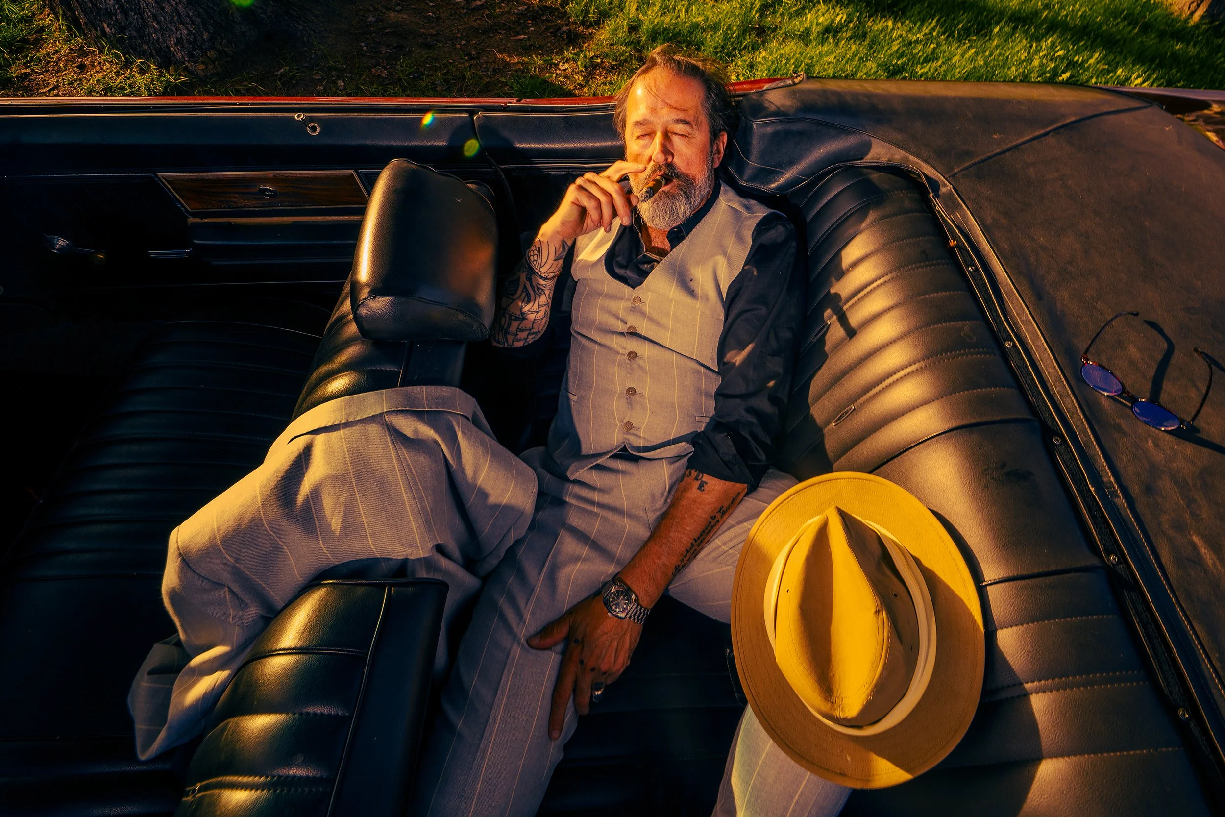 A man with a beard and tattoos sitting inside a vintage car, smoking a cigar. He is wearing a dark-colored shirt and beige plaid pants, with sunglasses resting on a yellow hat nearby. The scene is bathed in warm, golden light.