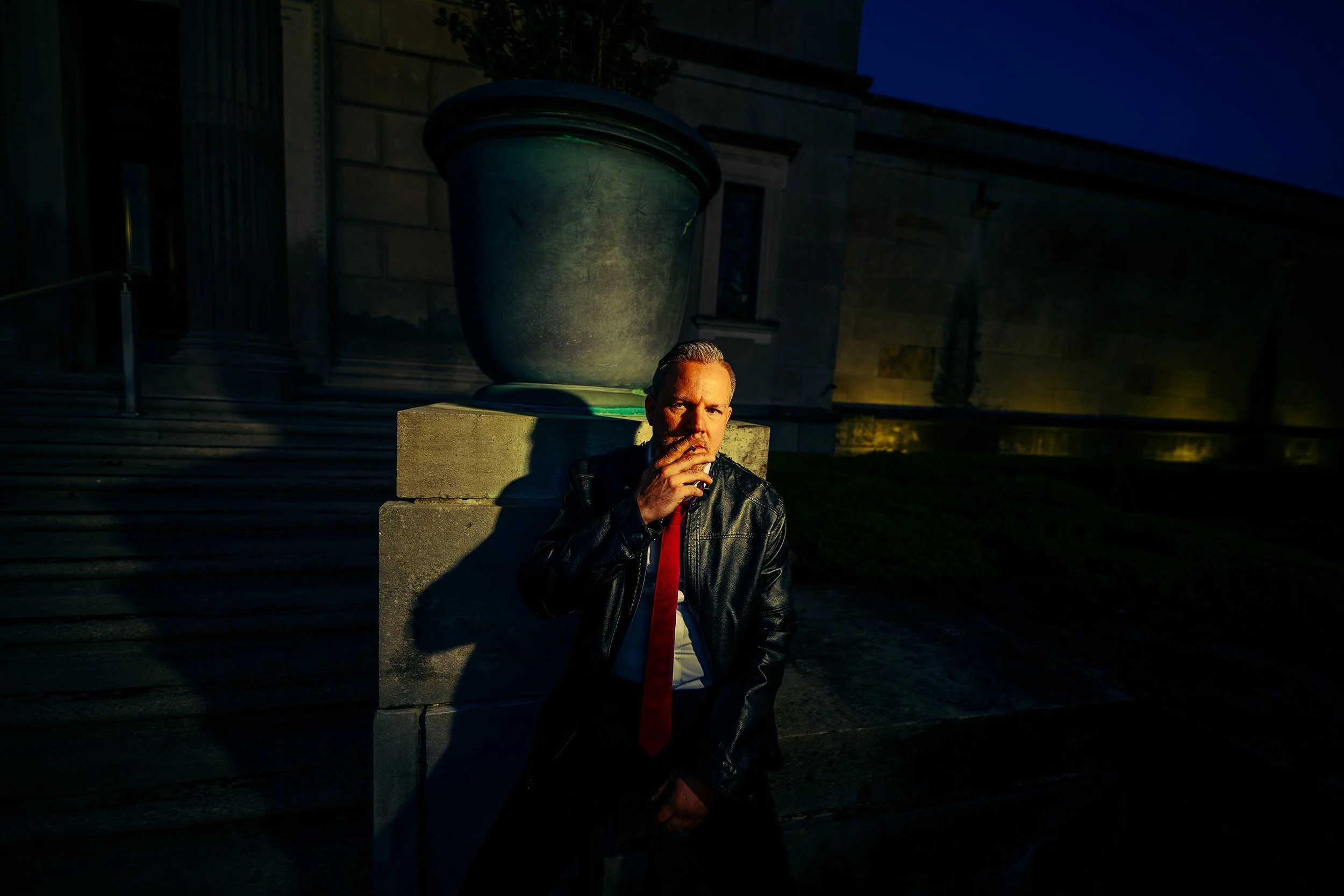 A man with slicked-back hair wearing a black leather jacket and red tie, sitting on a concrete bench outside at night, with a large greenish planter behind him. The background features a building with stairs and a dark evening sky.