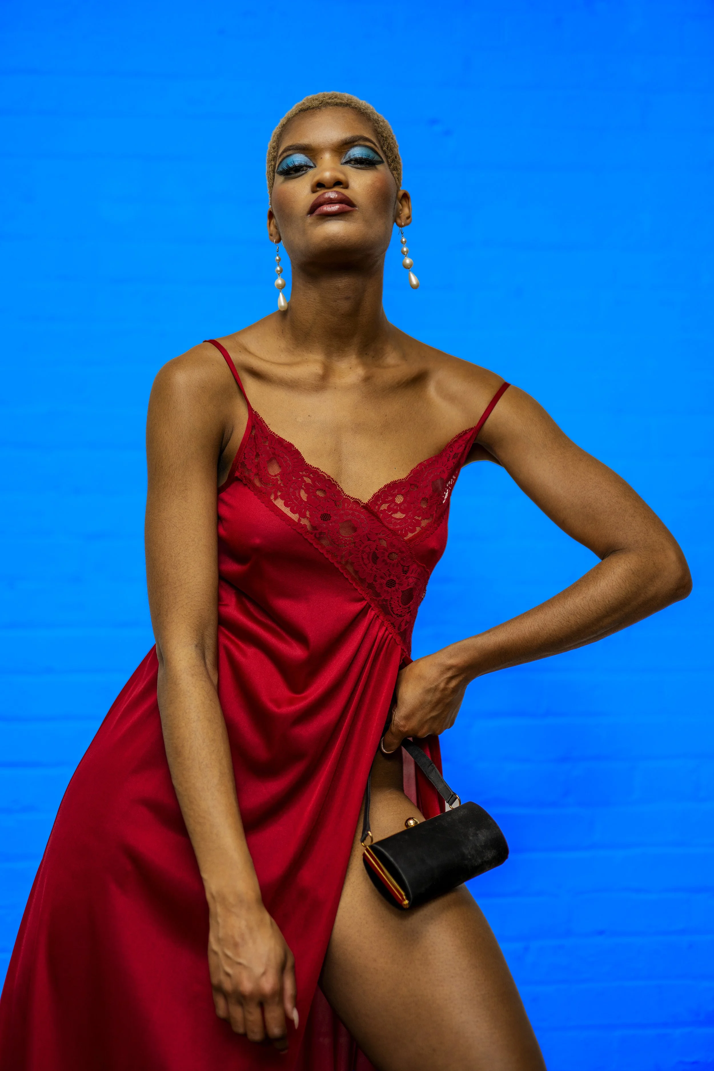A woman with short blonde hair and blue eye makeup wearing a red lace dress, holding a small black handbag with a gold clasp, standing against a bright blue background.