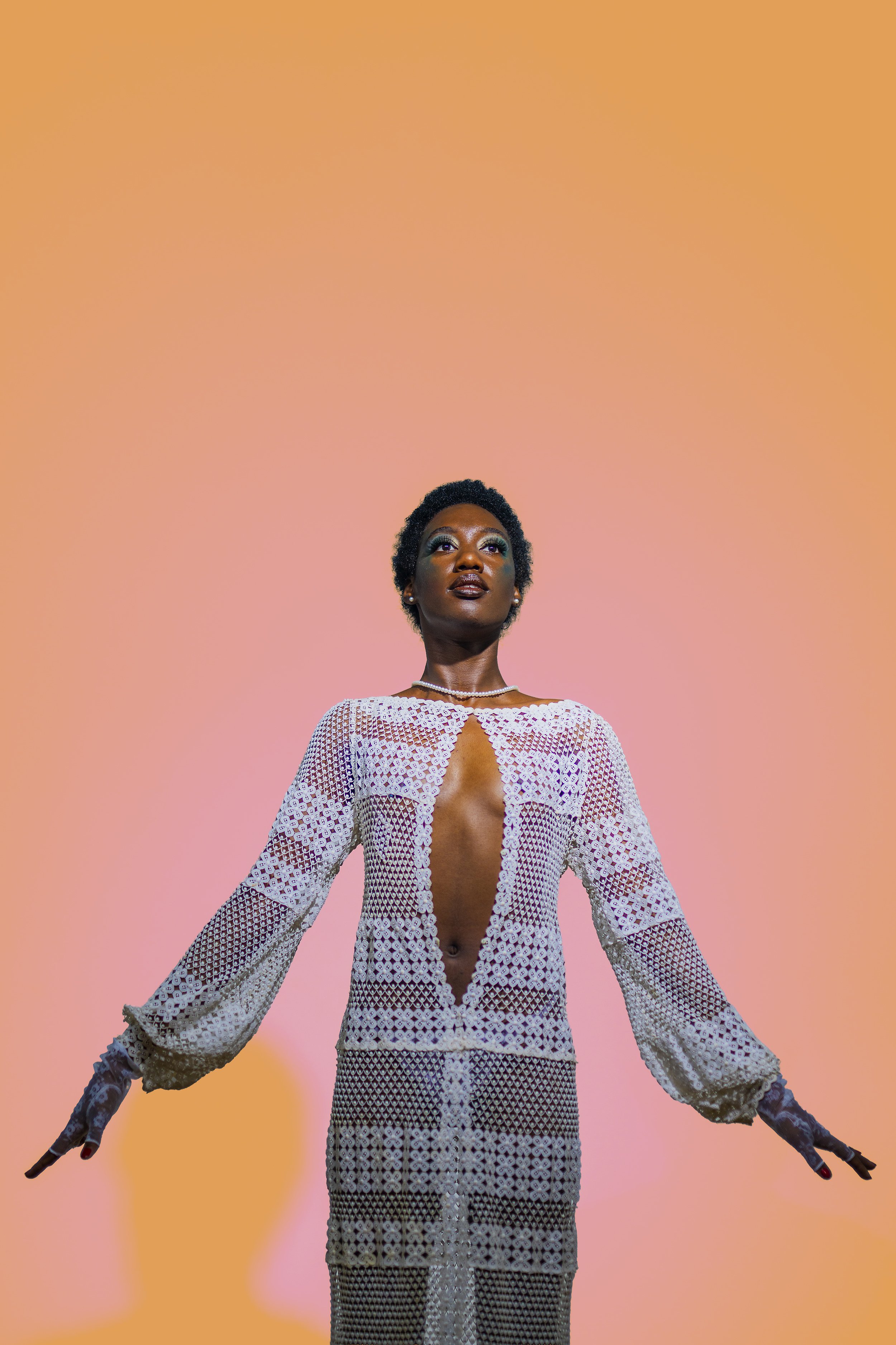 A woman with short curly hair and dark skin is standing against a pink and orange gradient background. She is wearing a white lace dress with a deep neckline and long sleeves. She is looking slightly upward with a serious expression.