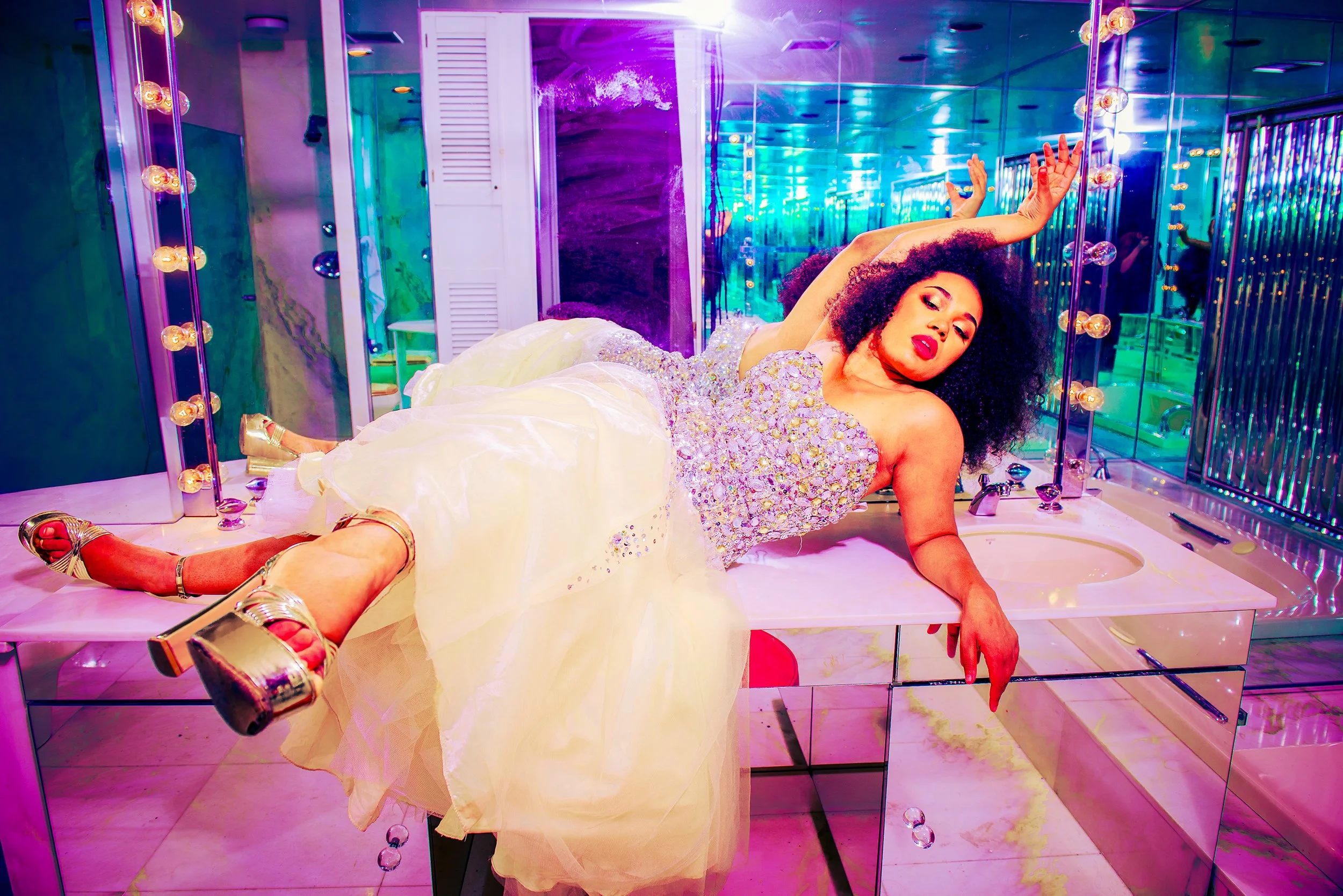 A woman with curly hair lying on a bathroom counter surrounded by mirrors and colorful lights, wearing a sparkling yellow dress and metallic high heels, with her arm extended and makeup makeup visible.
