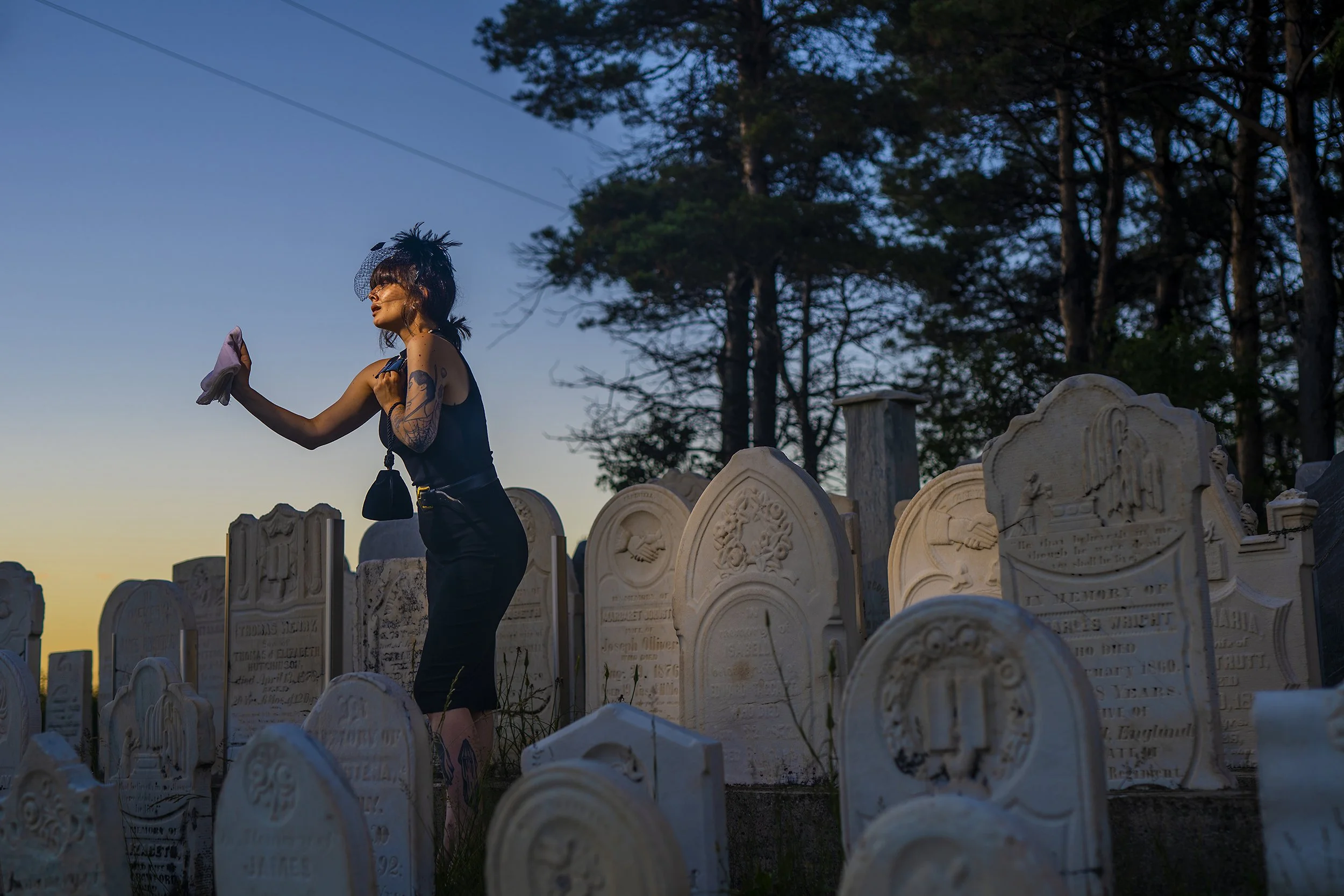 A woman with tattoos and dark hair styled in a messy updo, wearing dark clothing and sunglasses, stands among old tombstones in a cemetery at sunset, holding a cloth in one hand and a necklace in the other.