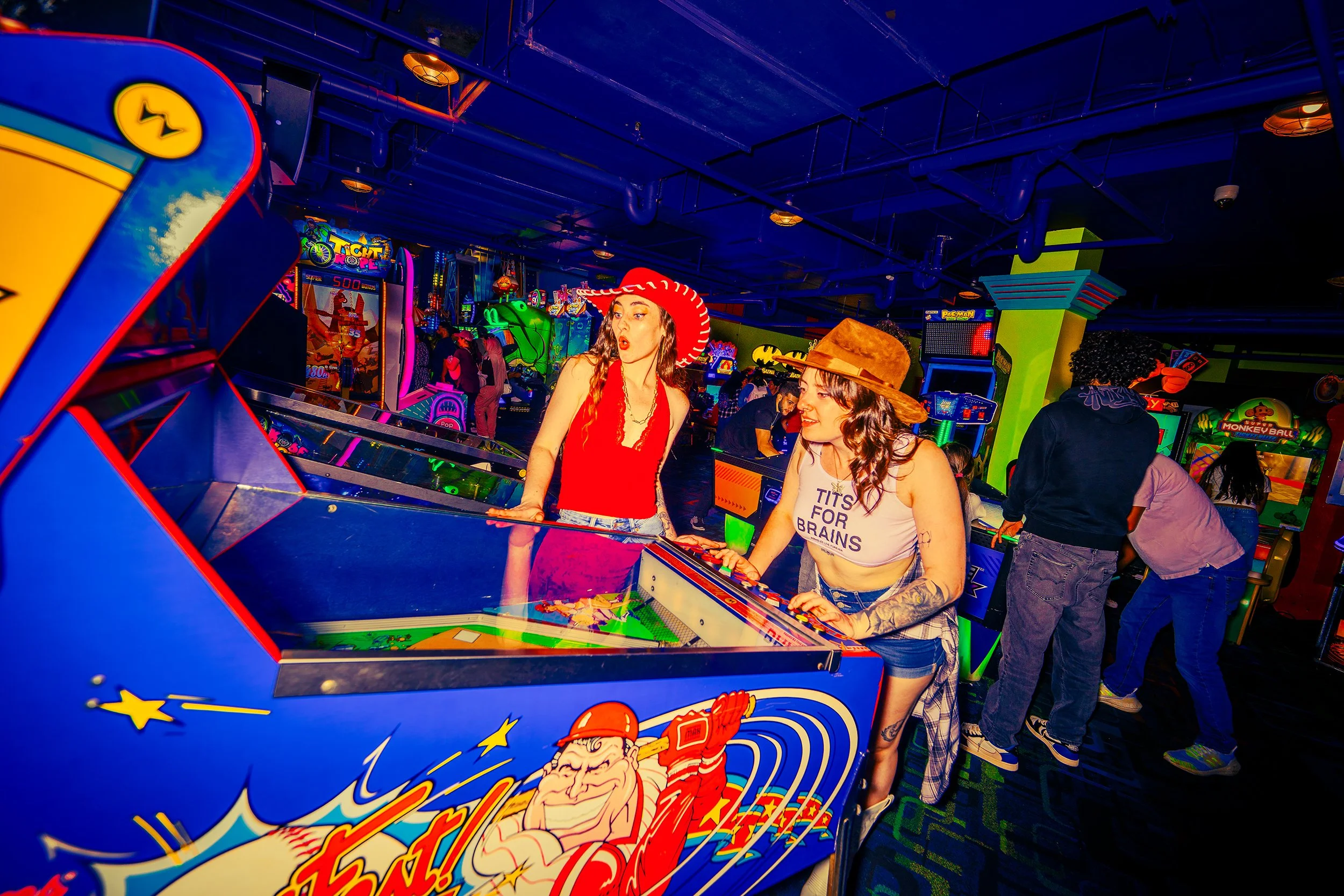 Two women playing an arcade game in a brightly lit arcade. The woman on the left wears a red hat and red outfit, while the woman on the right wears a brown hat and a white tank top with text. The arcade is filled with colorful game machines and other