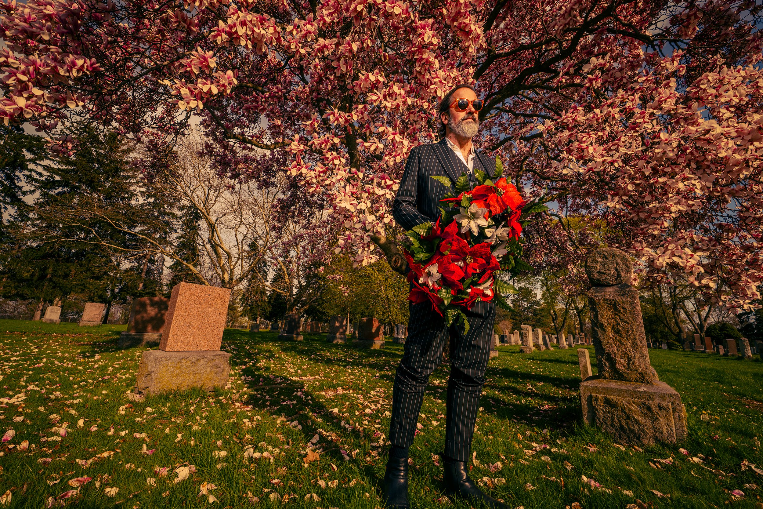 A man with sunglasses and a beard wearing a pinstripe suit holding a large bouquet of flowers standing in a graveyard with pink flowering trees in the background