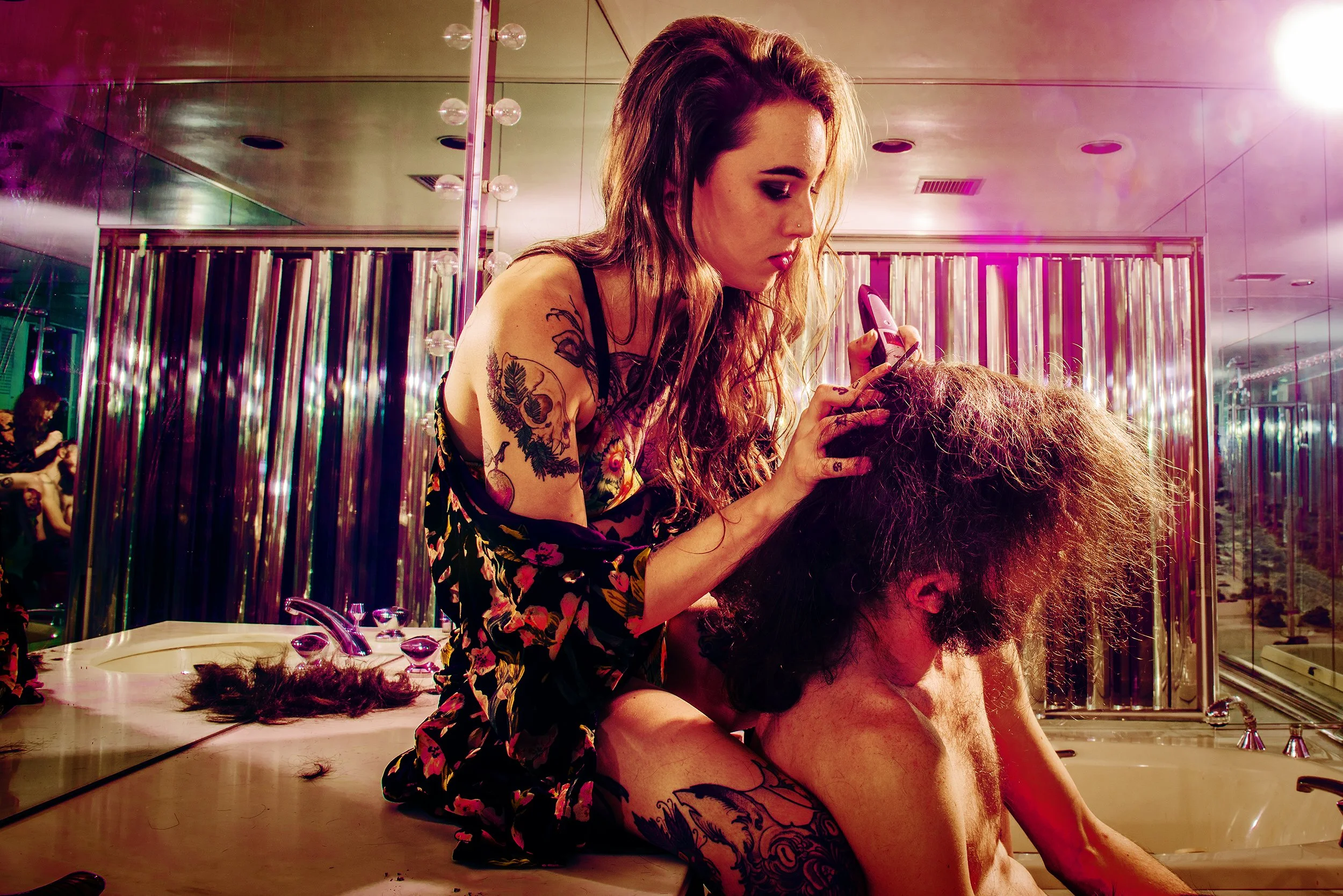 A hairstylist with tattoos on her arm styling a client's curly hair in a salon with pink and purple lighting.